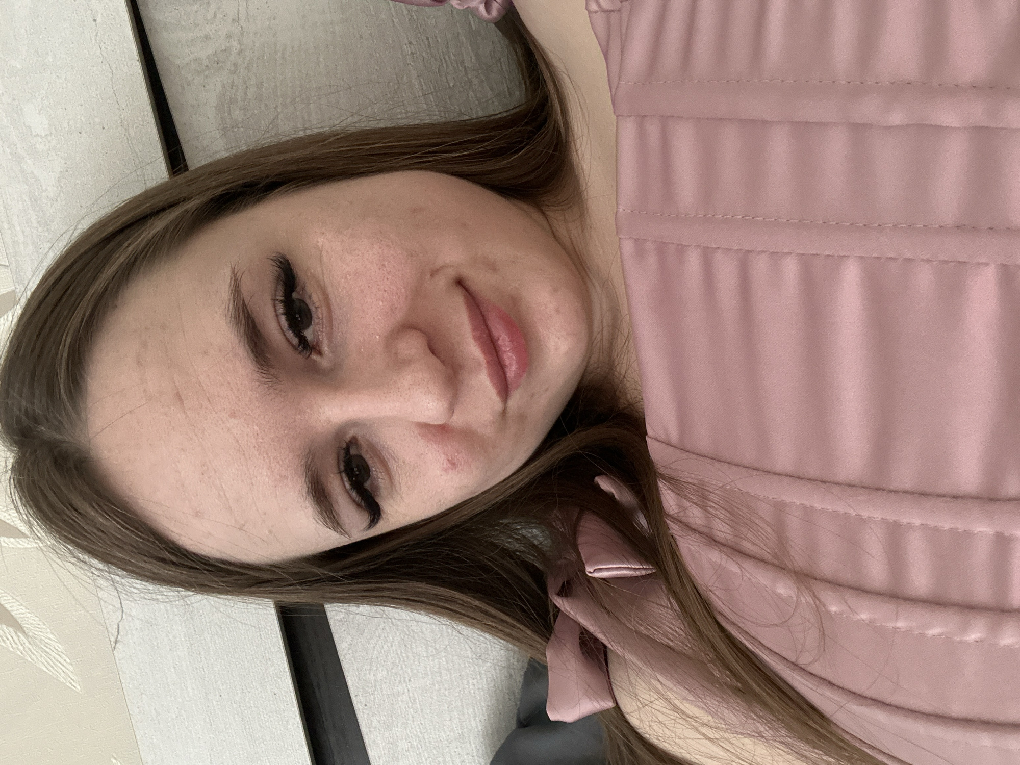 A woman is lying down. She has long brown hair and is wearing a pink top. She has long eyelashes. The background is a light-colored surface.
