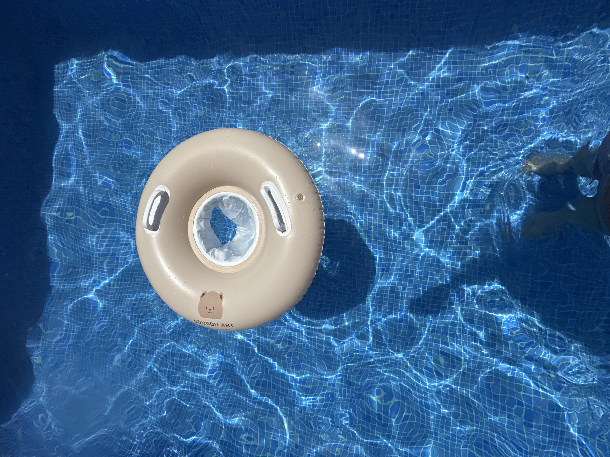 A tan-colored inflatable pool float with a white inner tube rests in the clear blue water of a swimming pool. The float has a cartoon bear graphic and the text "DUDDU ART" printed on its surface. The water is illuminated by sunlight, creating shimmering patterns of light and shadow on the tiled pool bottom. In the upper right corner, a person's legs and feet are visible, submerged in the water, suggesting they are either sitting in the pool or wading. The scene conveys a sense of relaxation and enjoyment on a bright day, likely in a warm climate, consistent with the location context of Sant Feliu De Guíxols, Spain, during daytime hours with clear weather. The overall impression is one of a leisurely summer day at a pool.