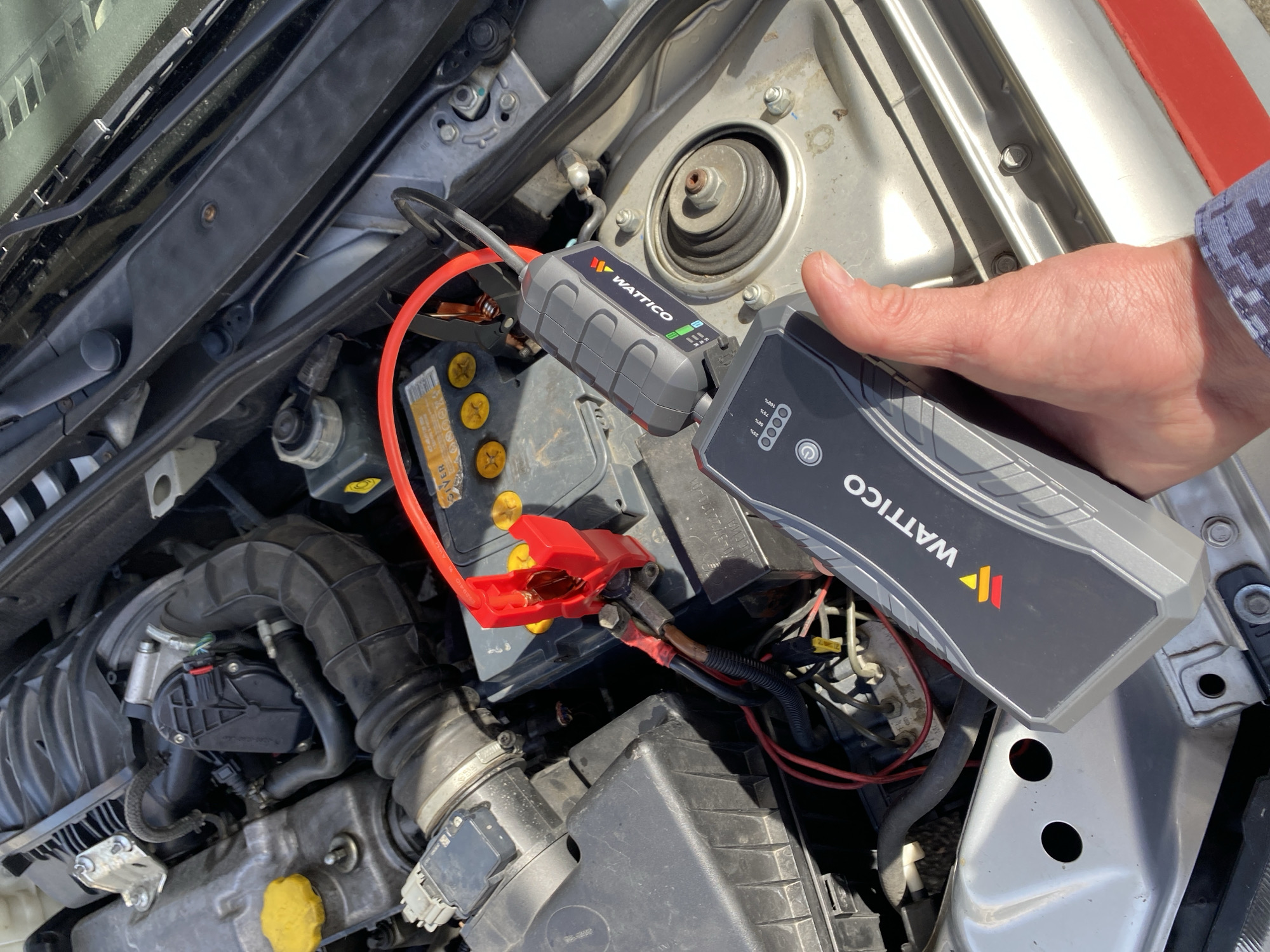 The image depicts a close-up view of a car's engine bay on a bright, sunny day in Sochi, Russia. A person's right hand, wearing a blue and white patterned shirt sleeve, holds a gray and black WATTICO branded jump starter pack.
The jump starter pack is connected via an adapter to the car's battery. The red positive clamp is visibly attached to the positive terminal of the car battery. The jump starter device displays LED indicators, with one green bar lit, and shows percentage markings for its charge level (0%, 25%, 50%, 75%, 100%). The car battery itself is a lead-acid type, appearing somewhat aged with yellow caps on its cells, and the word "EVER" is partially visible on its label.
The surrounding engine compartment reveals various components including black plastic air intake hoses, a silver engine block, fluid reservoirs, and wiring. The car appears to be an older model. The activity shown is the preparation or execution of jump-starting the vehicle, indicating a discharged car battery. The strong daylight suggests it is daytime.