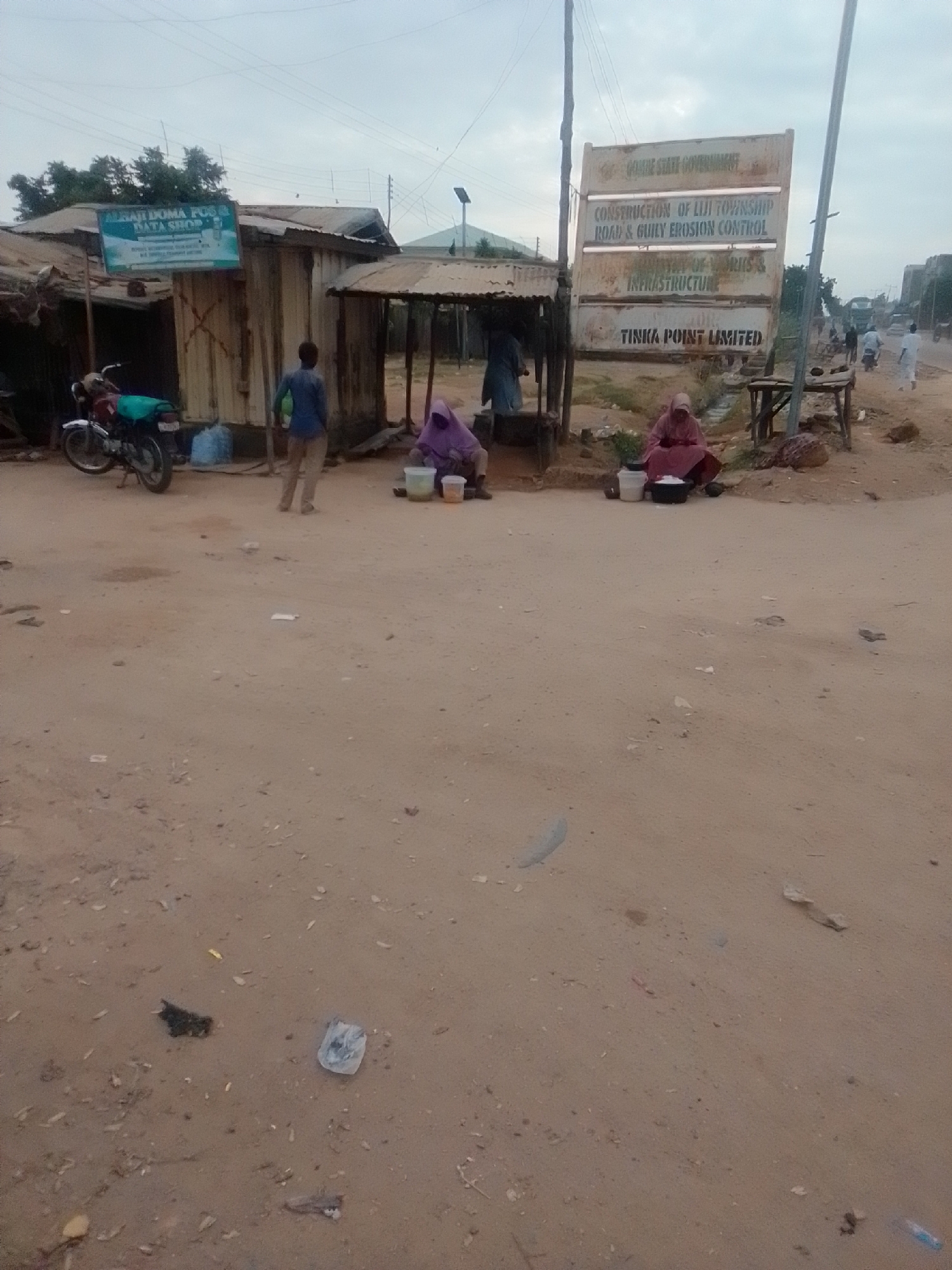 The image depicts a roadside scene in Gombe State, Nigeria, likely on a dirt road with some paved sections visible in the distance. A prominent sign in the center reads "GOMBE STATE GOVERNMENT CONSTRUCTION OF LIJI TOWNSHIP ROAD & GULLY EROSION CONTROL MINISTRY OF WORKS & INFRASTRUCTURE TINKA POINT LIMITED". Adjacent to the sign is a small stall or shop with a sign that reads "ALHAJI DOMA POS & DATA SHOP".
In the foreground, the ground is mostly dirt and sand, with scattered debris including plastic bags and other refuse. To the left, a motorcycle is parked in front of a structure. A person dressed in blue is standing near the shop, and another individual in a purple headscarf is seated on the ground, seemingly selling items from buckets. Further back, a person in a pink headscarf is also seated, with containers in front of them. In the background, traffic, including motorcycles and vehicles, can be seen on the road, and there are buildings and vegetation. The sky is overcast, suggesting an early morning or late afternoon setting. The overall impression is a busy, functional area on the outskirts of a town or city.