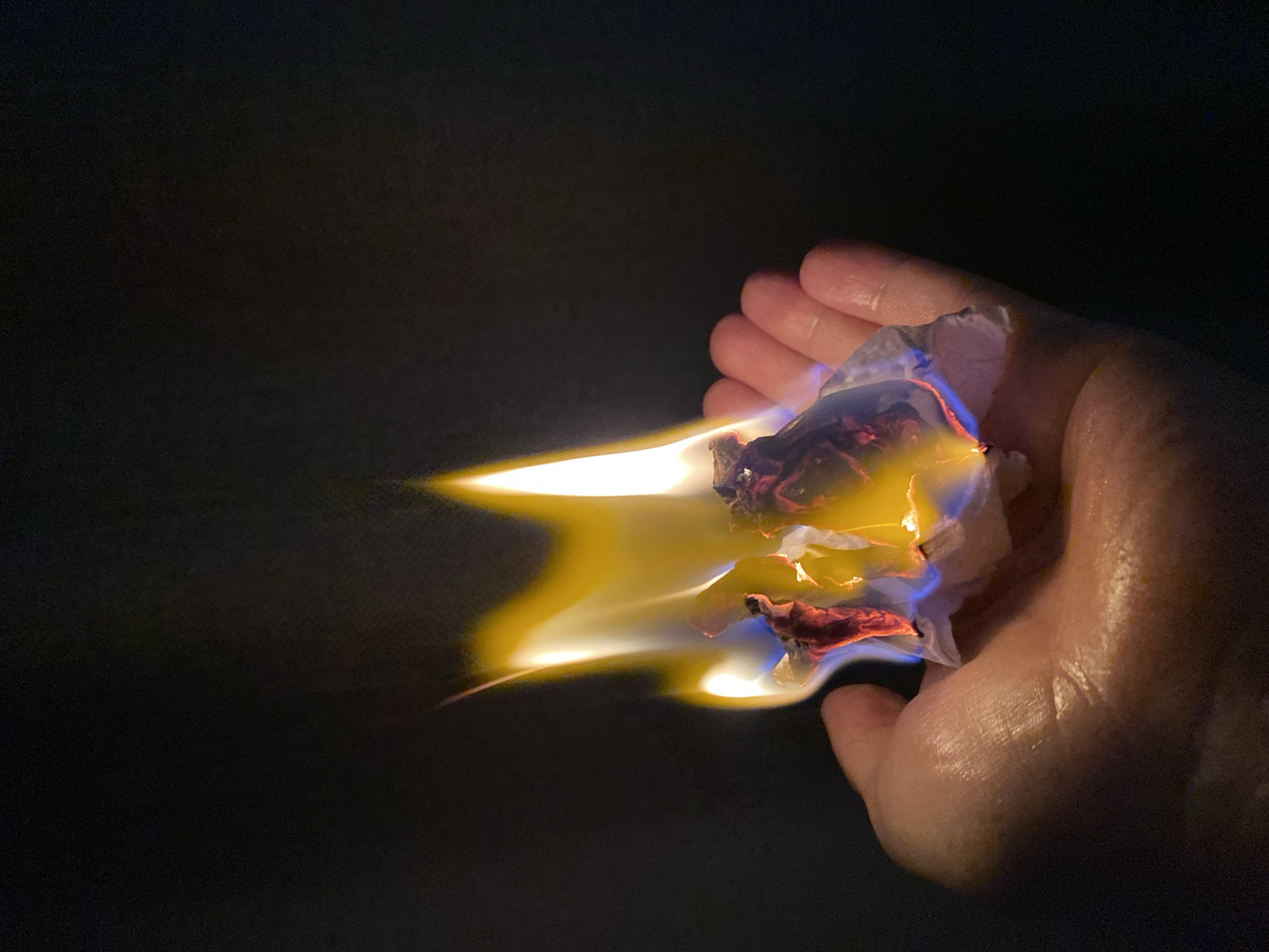 A hand holds burning material in a dark environment. The fire is yellow and blue, and remnants of red material are visible within the burning object. The hand is Caucasian and appears slightly moist. The location is Unknown, Unknown.