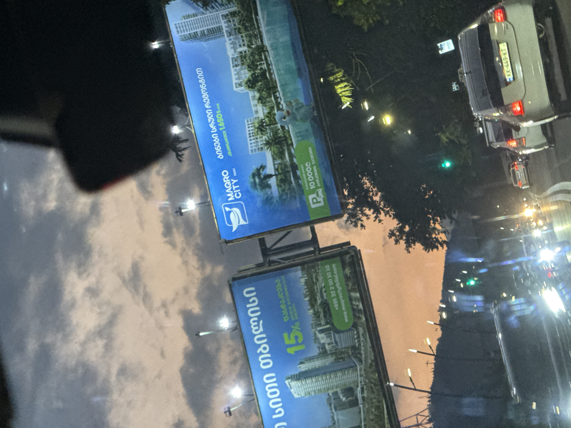 The image captures a view from a vehicle driving through the city of Tbilisi, Georgia, likely during the evening or twilight. Two prominent billboards are visible, advertising a real estate development called "Magro City". The left billboard features an image of modern high-rise buildings and text in Georgian, along with a "15%" discount offer. The right billboard, also featuring Georgian text, displays "Magro City" and an image of a swimming pool area with a cartoonish blue bird. It also shows "1,650₾" and a parking icon with "10,000₾".
The environment suggests a road with multiple lanes of traffic, indicated by the blurred motion of other vehicles and streetlights. The sky is cloudy with hues of orange and grey, typical of sunset or sunrise. The lighting from streetlights and vehicle headlights creates a somewhat dramatic and atmospheric scene. The reflection on the vehicle's windows suggests the photograph was taken from inside another car. The license plate of the white car in the foreground reads "KK-449-KT".