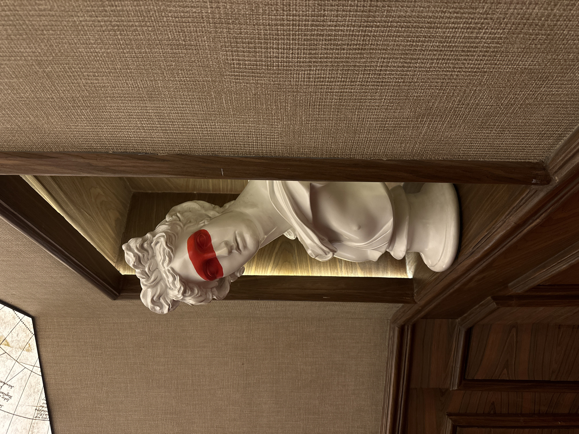 A white bust with a red band over its eyes sits in a recessed shelf with warm lighting. The shelf is framed by dark wood and is positioned beneath a tan textured ceiling. Adjacent to the shelf, a map hangs on the wall. The overall scene suggests an interior space with decorative elements. There is no indication of the location, Itirin, Nigeria.