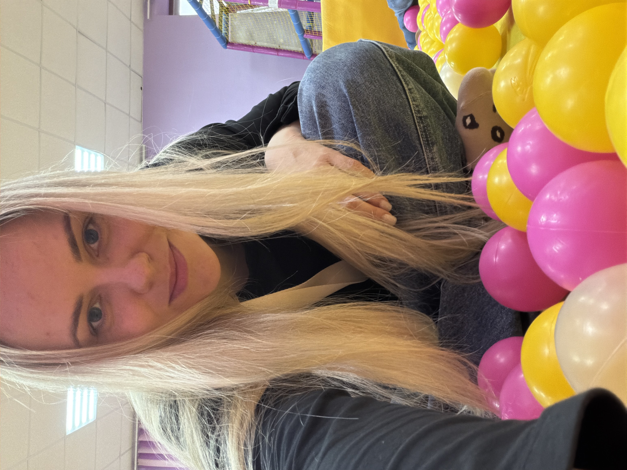 A blonde woman is featured in an indoor setting, likely a play area or entertainment center in Votkinsk, Russia. She is partially submerged in a ball pit filled with vibrant pink and yellow plastic balls. The woman, with long, straight blonde hair fanned out around her, looks directly at the camera with a subtle smile, conveying a sense of relaxed enjoyment. She is wearing a black long-sleeved top and blue jeans. Her left hand is visible, resting near her leg, and a beige sock with a dark, abstract pattern is seen on her foot, peeking out from the balls.
The background of the image reveals elements of an indoor facility designed for recreation. A purple wall is visible behind her, contrasting with a white ceiling featuring grid patterns and bright fluorescent lighting. Part of a colorful play structure with blue and yellow components is also discernible, reinforcing the setting of a children's play area or similar recreational venue. The bright colors of the balls and the overall environment suggest a cheerful and playful atmosphere. No visible text is present in the image.
