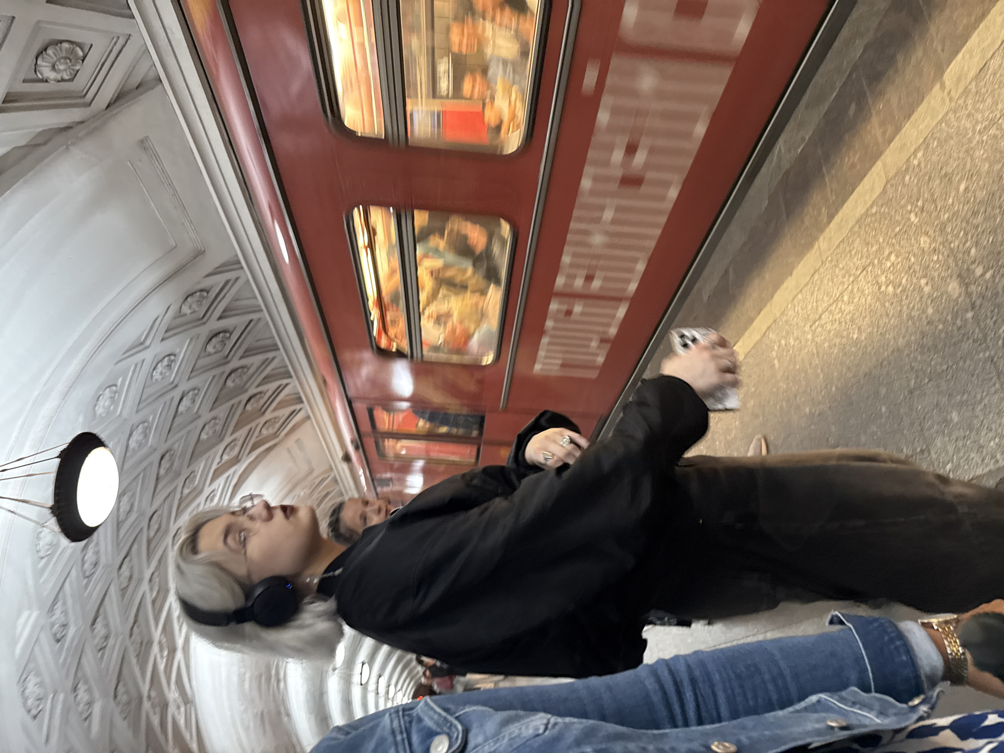 An eye-level, medium shot shows a young woman wearing headphones and a black jacket, standing on a subway platform in Moscow, Russia, looking to her right. The woman has short, blonde hair and is wearing glasses. To her left, a person wearing a denim jacket is partially visible. A red subway train with blurred windows is passing behind her, and passengers are visible through the windows. The subway station has a decorative vaulted ceiling with ornate patterns and a hanging light fixture. The platform floor is a textured grey stone with a yellow safety line. The woman is holding a smartphone in her right hand.
