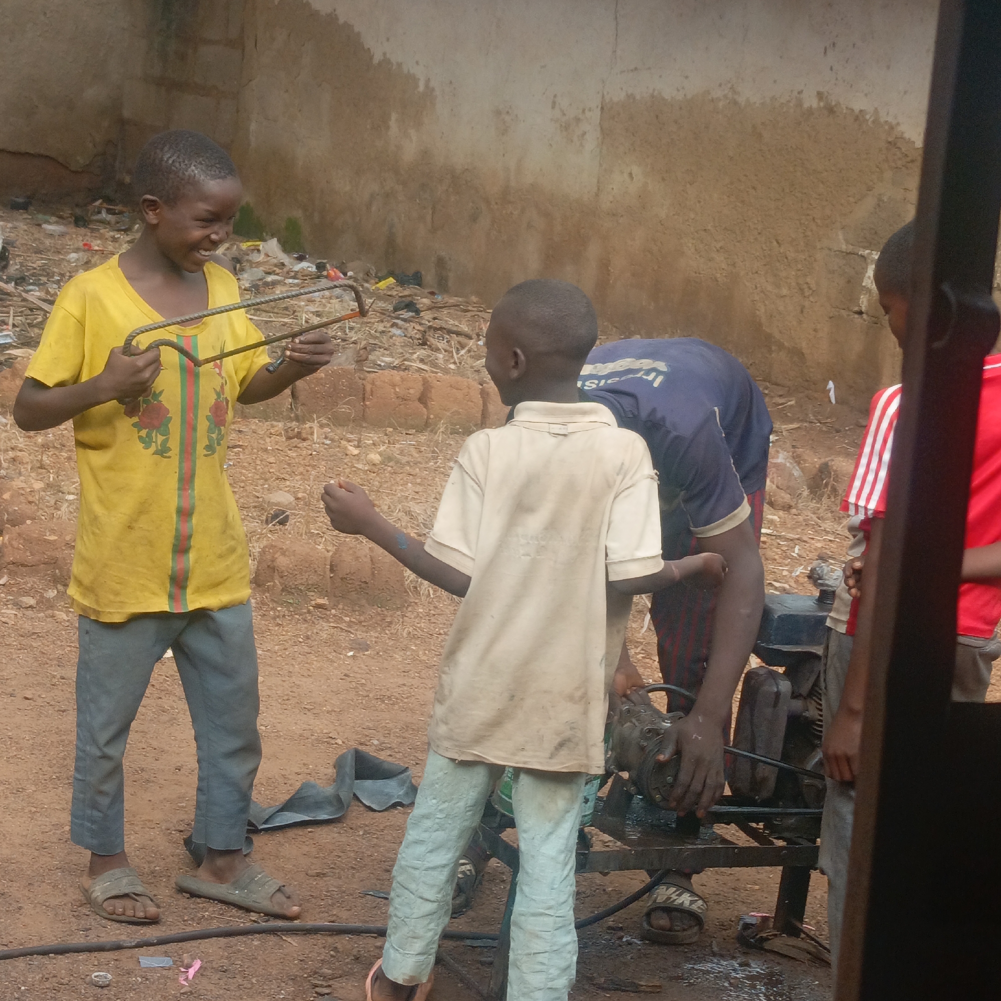 In a dusty, outdoor setting, possibly in Akwanga, Nigeria, four young boys are engaged in various activities around a metal object. The boy on the left, wearing a yellow t-shirt with red and green stripes and an emblem of red flowers, holds two bent pieces of rebar, looking towards the boy opposite him with a wide, toothy grin. The boy in the center, wearing a light-colored t-shirt and light blue pants, faces the boy in yellow, with his right arm extended. Another boy, wearing a dark blue t-shirt with white lettering and red and black striped pants, is bent over, with his hands on a metal contraption that appears to be a generator or engine. A fourth boy, partially visible on the far right wearing a red and white striped shirt, stands near the metal object. The ground is composed of reddish-brown soil, scattered with debris including plastic bags and what looks like a deflated inner tube. The background features a rough, plastered wall with a brick foundation. The overall impression is one of children interacting and working in a semi-industrial or backyard environment.