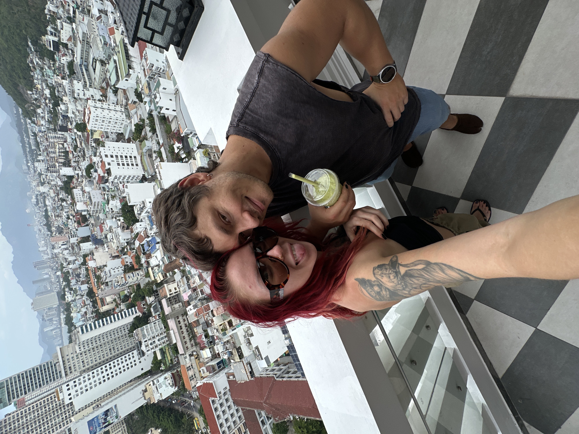 A couple, a woman with bright red hair and a man with short brown hair, stand on a balcony overlooking the city of Nha Trang, Vietnam. The woman, wearing large sunglasses and a black top, sticks her tongue out at the camera. She has a tattoo of an angel on her left arm. The man, wearing a dark grey sleeveless shirt and denim shorts, holds a green smoothie. They are positioned in the foreground, with the sprawling city, featuring numerous buildings and distant mountains, spread out behind them. The balcony floor is tiled in a black and white checkered pattern.