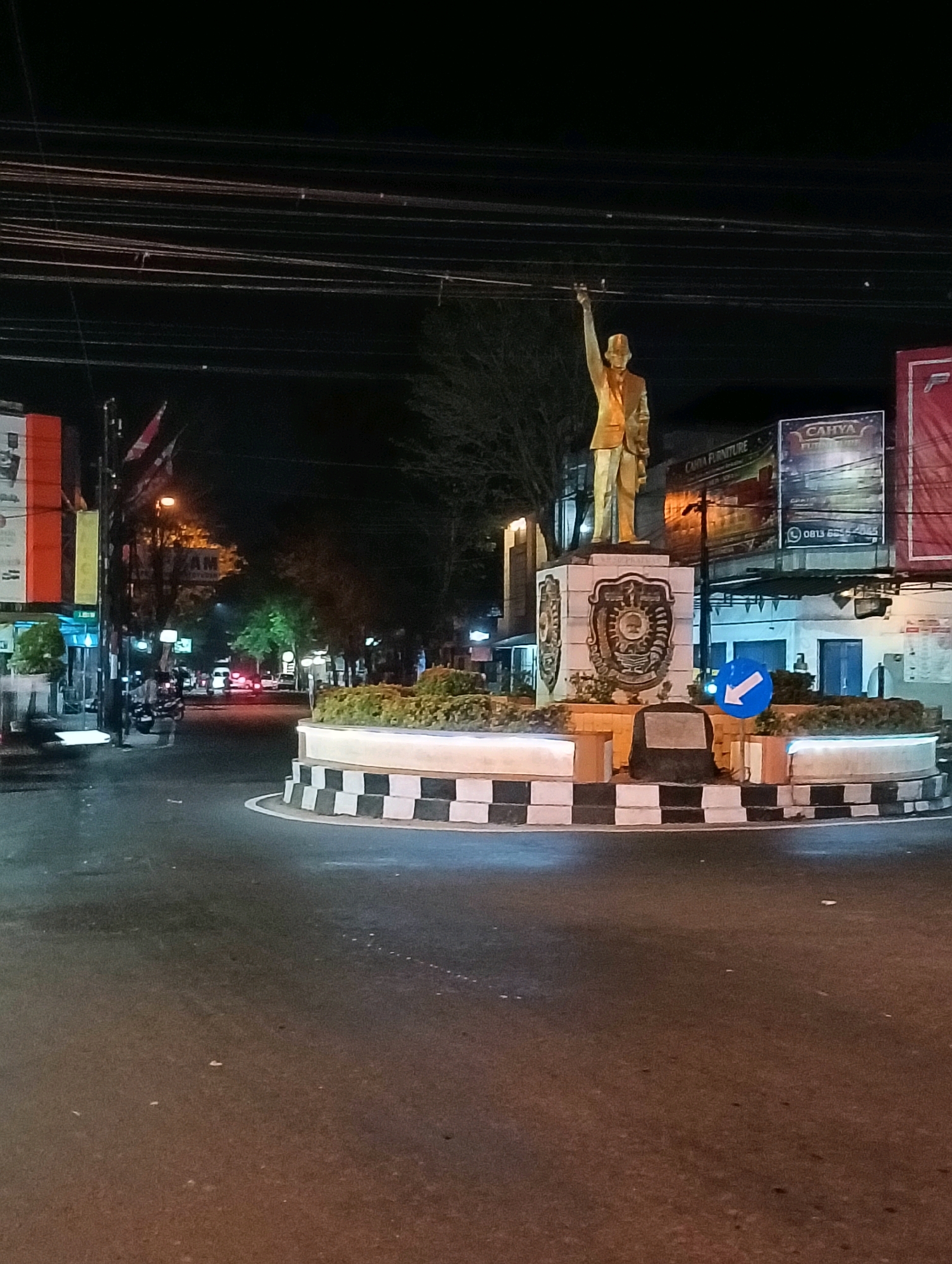 The image shows a roundabout at night in Purworejo, Indonesia. A golden statue of a man in a suit with his arm raised stands in the center of the roundabout on a decorated pedestal. The roundabout is bordered by black and white checkered patterns. In the background, there are trees, buildings, streetlights, and overhead power lines. A blue traffic sign with a white arrow pointing to the right is visible on the side of the roundabout. A few vehicles are seen in the distance on the road. The scene is illuminated by artificial lighting, creating a nocturnal ambiance.