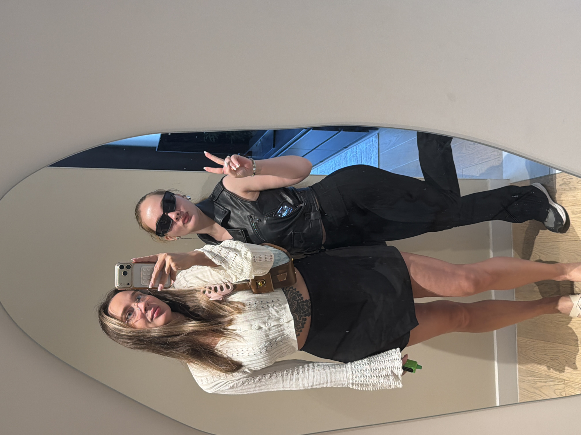 Two young women are posing for a mirror selfie in a room that appears to be a dressing room or entryway. The woman on the left has long, light brown hair and is wearing glasses, a white cropped long-sleeved top with bell sleeves, and a black skirt. She is holding a smartphone to take the picture. She also has a tattoo visible on her stomach. The woman on the right has her hair pulled back and is wearing dark sunglasses, a black sleeveless leather vest over a dark top, and black pants. She is making a peace sign with her right hand. The background shows a modern, minimalist interior with light-colored walls and a wooden floor. The mirror they are using is large and has an irregular shape. There are cabinets visible in the reflection of the mirror behind the women. The overall impression is that of a fashionable and casual moment captured indoors. There is no specific indication of a location in Moscow, Russia.