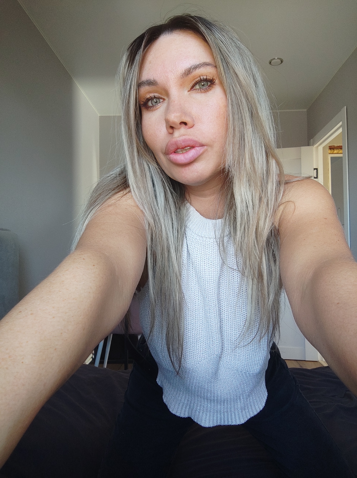 A light-skinned woman with blonde hair is taking a selfie. She is wearing a white knit top and black pants. The background appears to be a bedroom with gray walls and a partially visible door. The location is not discernible as being Votkinsk, Russia.