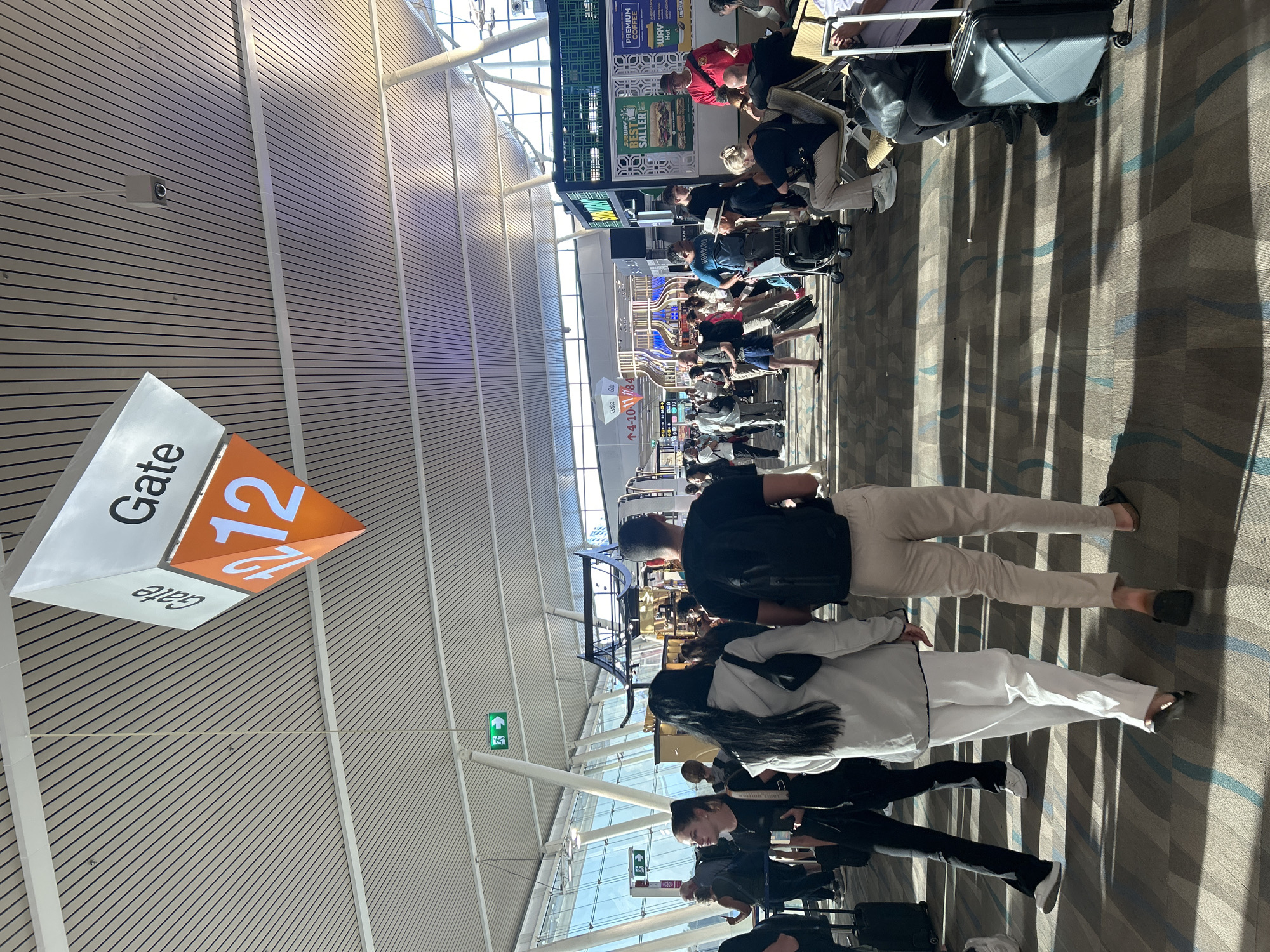 The image depicts an interior scene at an airport terminal. A prominent sign hanging from the ceiling indicates "Gate 12". In the foreground, several individuals are walking and standing, some with luggage. One woman in the foreground is wearing a white jacket and white pants, while a man next to her is wearing a black t-shirt and light-colored pants. Behind them, more people are visible, some queuing and others seated. The environment includes large windows allowing natural light to enter, a patterned carpeted floor, and a high ceiling with a distinctive slatted design. Signs with green directional arrows are also present on the walls. The overall impression is that of a busy airport gate area.