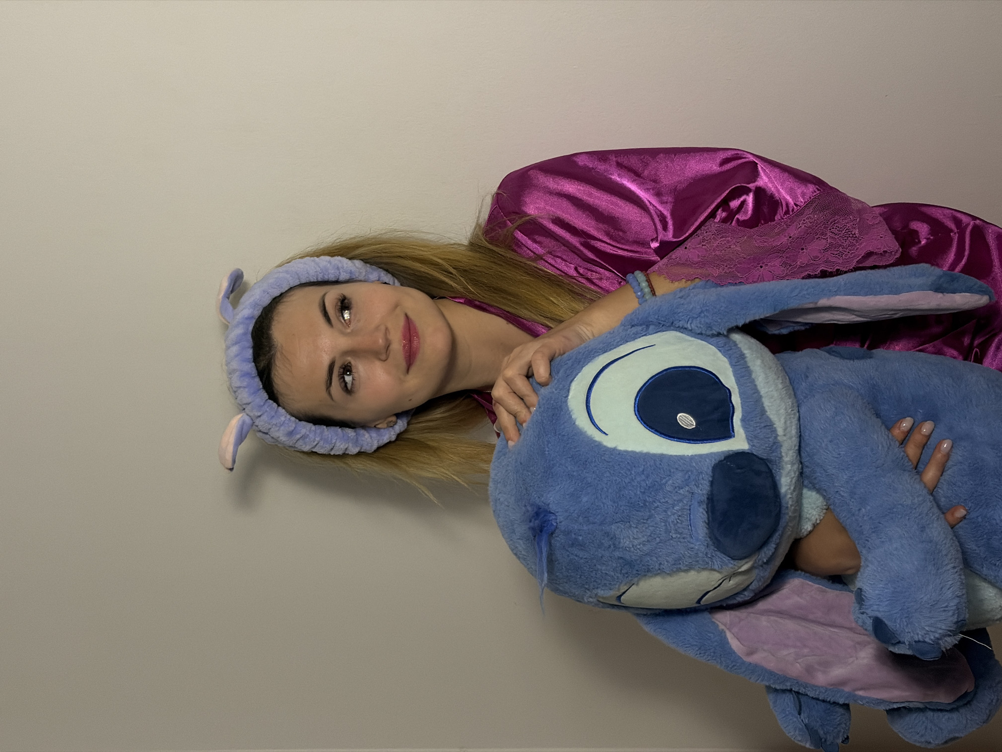 A woman in a purple robe and headband is lying down and holding a large blue stuffed animal that resembles Stitch from Disney's Lilo & Stitch. The location is not discernible from the image.