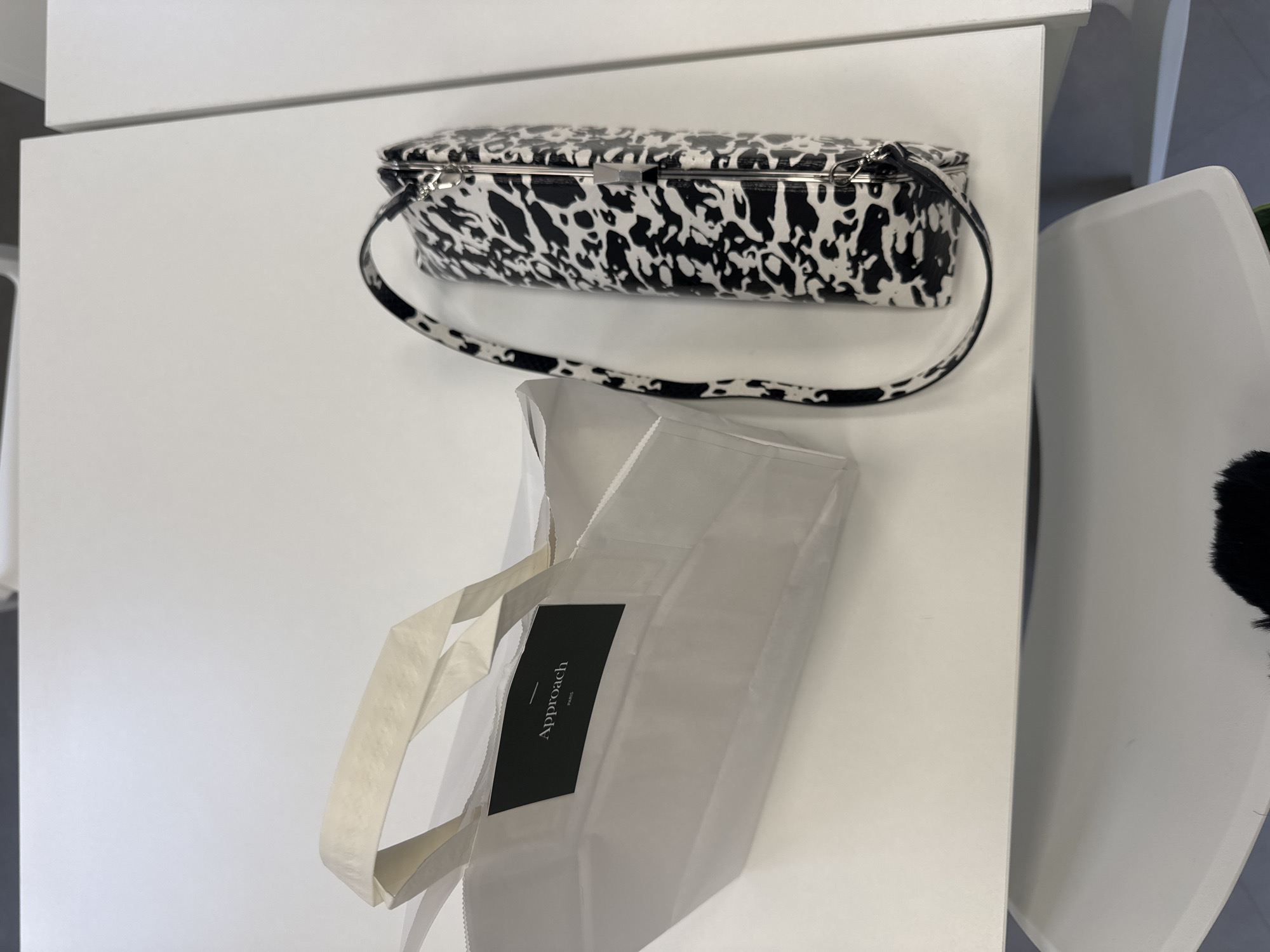 A high-angle medium shot depicts a white handbag patterned with black spots and an off-white paper shopping bag labeled "Approach PARIS" in black. The bag sits on a white surface, with a white chair visible in the background, suggesting an interior setting. The scene suggests a recent purchase from a Paris-based retailer. No people are visible.