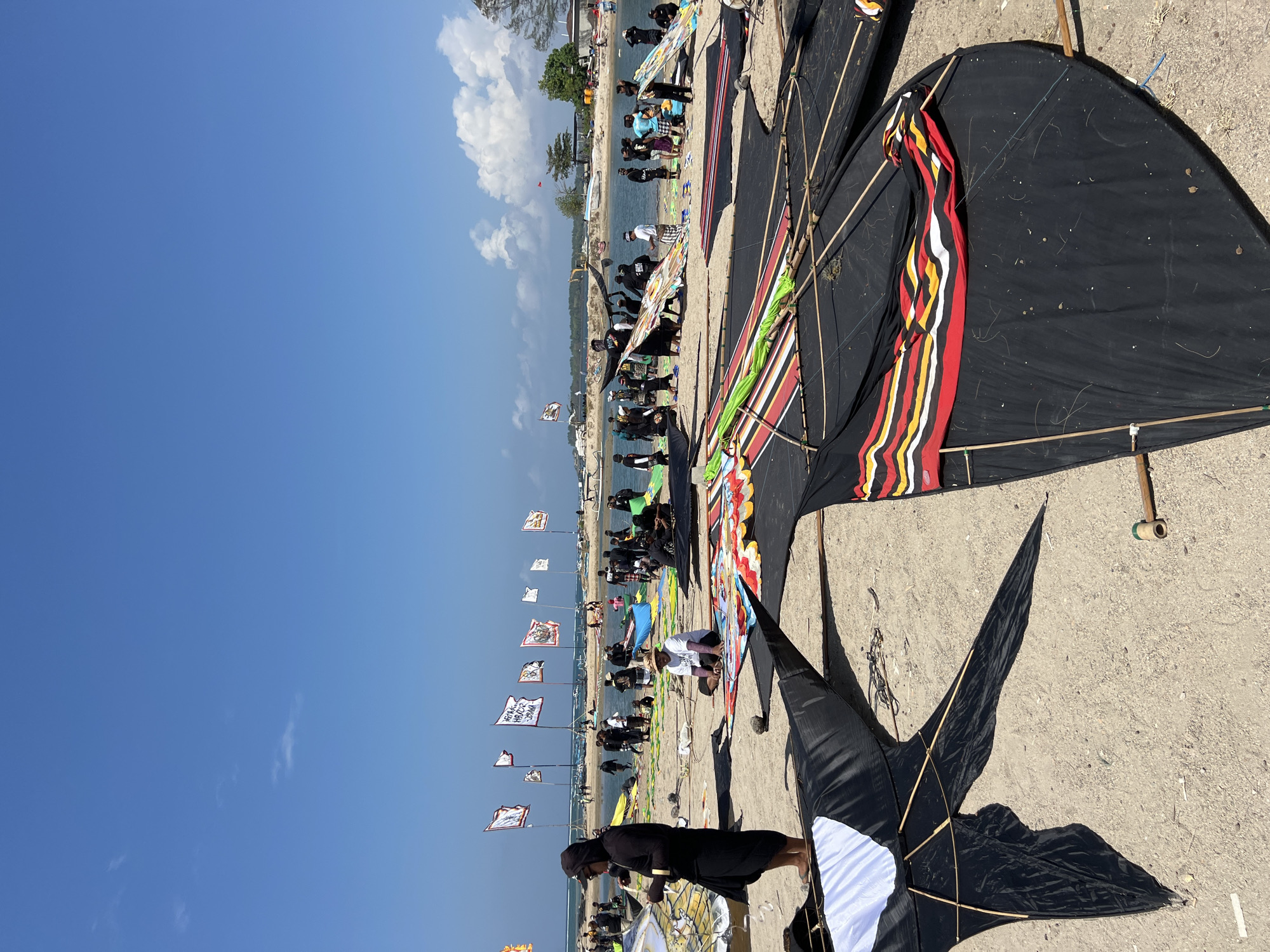 This image depicts a beach scene in Denpasar, Indonesia, likely during a kite festival or preparation for one. The foreground is dominated by several large, traditional kites laid out on the sand, predominantly black with colorful decorative patterns, primarily red, yellow, and white stripes. Some kites are partially assembled with visible bamboo frames.
In the mid-ground, numerous people are gathered on the sandy beach and by the water's edge. Many are dressed in dark clothing, including head coverings, suggesting a local cultural event. Several large flags with various symbols and text are planted in the sand along the shoreline. These flags are a prominent visual element, adding to the festive atmosphere.
The background shows a calm body of water, likely the ocean, with a distant shoreline featuring vegetation and some structures. The sky is clear blue with a few scattered white clouds. The overall scene conveys a sense of community gathering and preparation for a cultural activity on a sunny day.