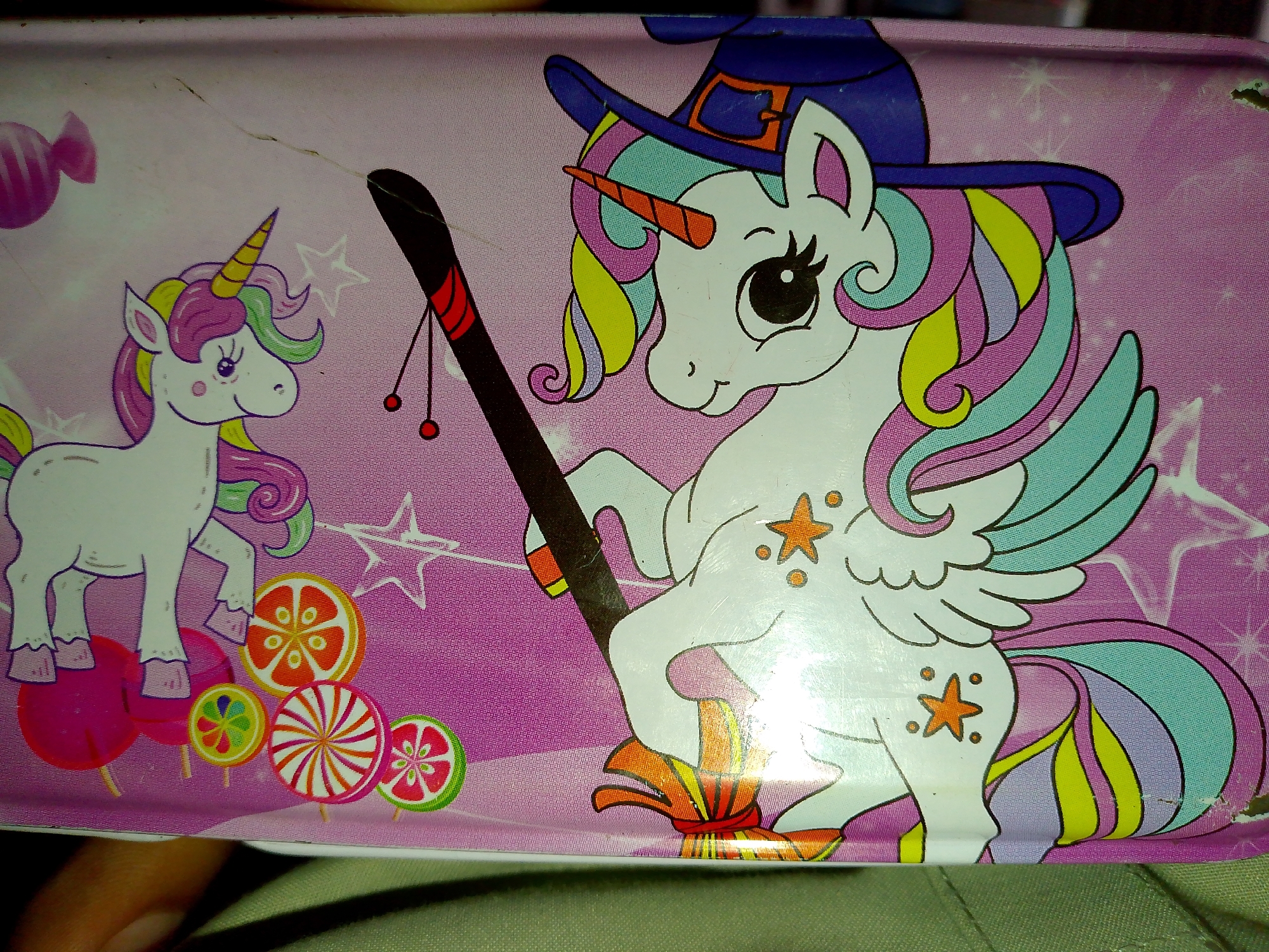 The image shows a close-up of a pink, patterned tin. The tin features illustrations of two unicorns and various candies. The unicorn on the left is white with a rainbow mane and tail, wearing a pink horn and standing on a pink circular object. To its left is a pink candy with stripes. In the foreground, to the left and center, are several lollipops of different colors and patterns, including orange, yellow, red, and green. The unicorn on the right is also white with a rainbow mane and tail, wearing a blue witch's hat with an orange buckle. This unicorn is larger and positioned more centrally, holding a black broomstick with red and black accents. The broomstick has two small red balls hanging from it. The background is a light pink with scattered white stars. A human hand is visible at the bottom left corner of the image, partially obscuring the tin.