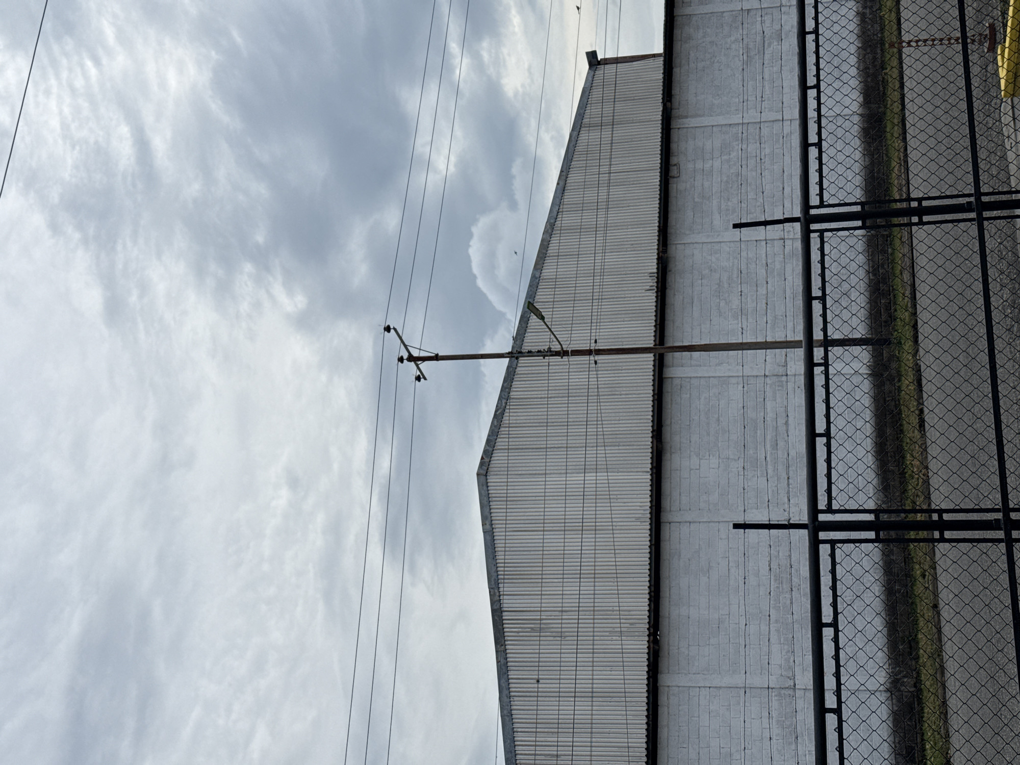 The image depicts an exterior scene in Tinaquillo, Venezuela, oriented sideways. A large, light-colored building with corrugated metal siding and a concrete base is prominent on the right. A section of chain-link fence runs along the right side of the building. Overhead power lines stretch across the cloudy sky on the left. A utility pole with insulators is visible against the sky. A horizontal rod is fixed to the wall of the building.