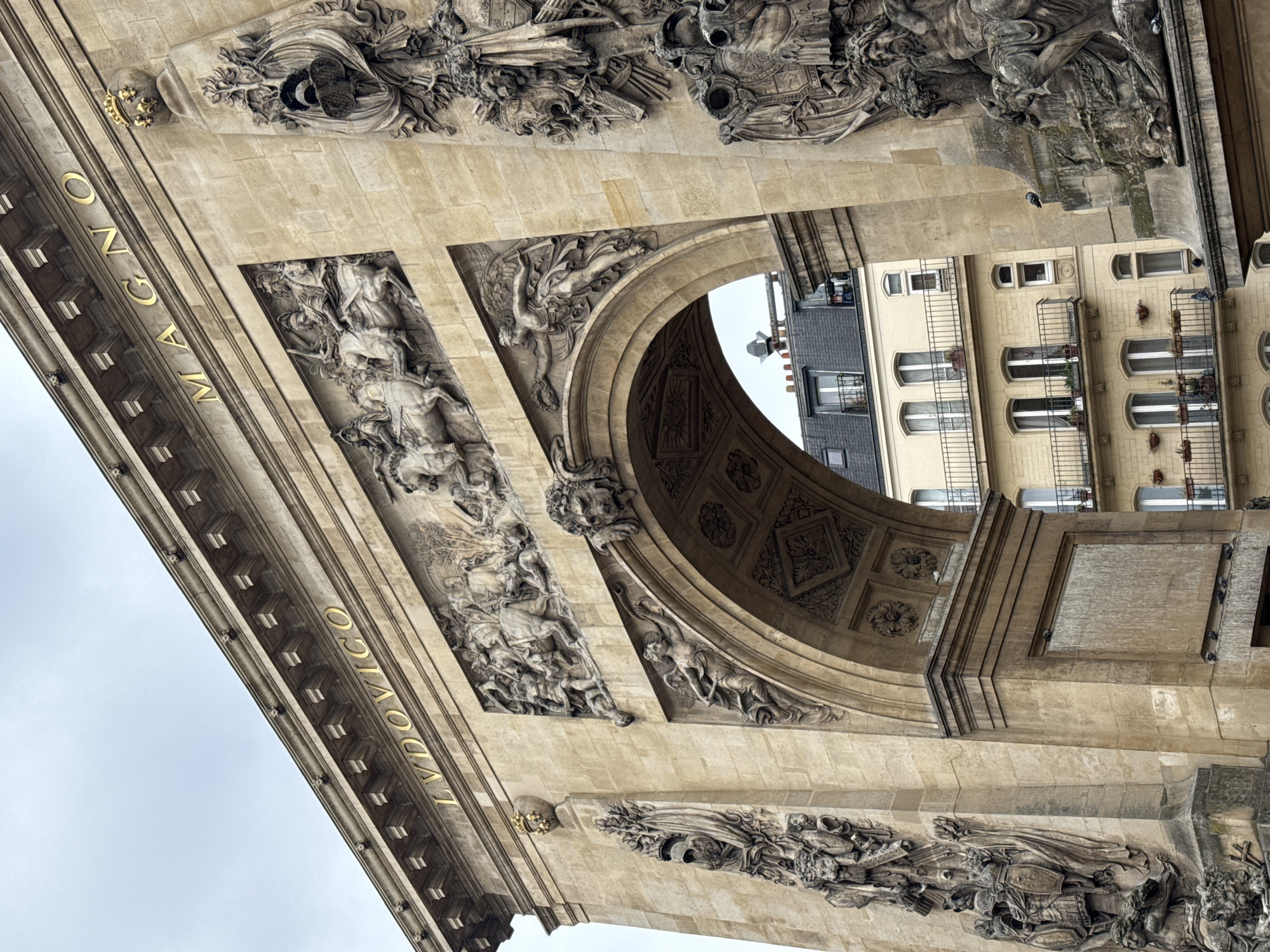 This image depicts a low-angle view of the Arc de Triomphe du Carrousel in Paris, France. The sandstone arch is highly decorated with bas-relief sculptures and inscriptions. The name "LUDOVICO MAGNO" is visible in gold lettering on the arch. Through the central archway, the facades of buildings across from the arch are visible.