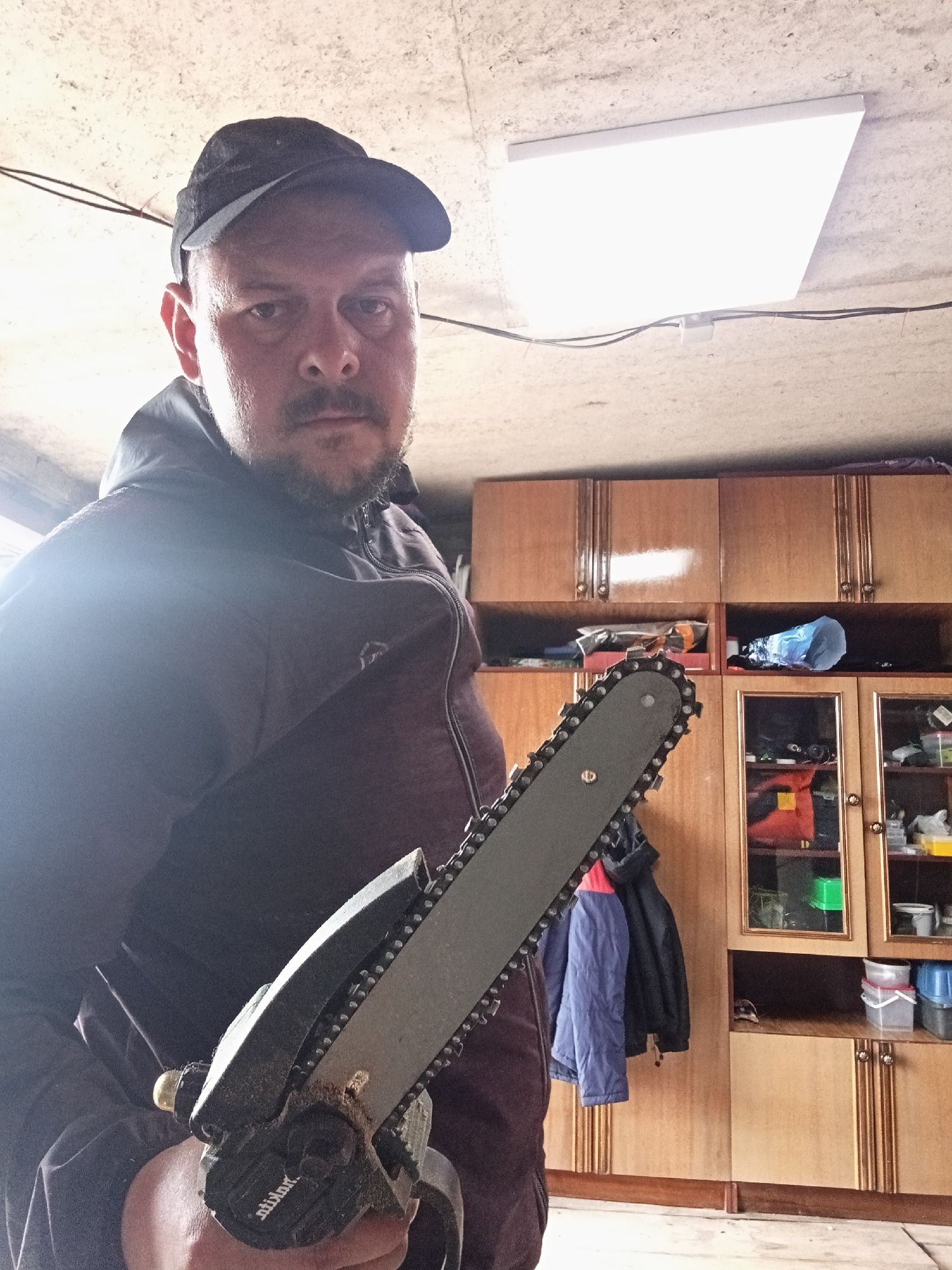 A man wearing a black baseball cap and a dark hooded jacket is holding a chainsaw. He is standing in what appears to be a garage or workshop, with wooden cabinets and shelves filled with various items in the background. The ceiling is concrete with a fluorescent light fixture. The man's face is partially obscured by the chainsaw, but he has a beard and mustache. The lighting is bright, creating a lens flare on the man's jacket. The image is taken from a low angle, looking up at the man. The scene is likely indoors, as indicated by the presence of cabinets and the artificial lighting. The overall impression is that of a casual or informal setting.