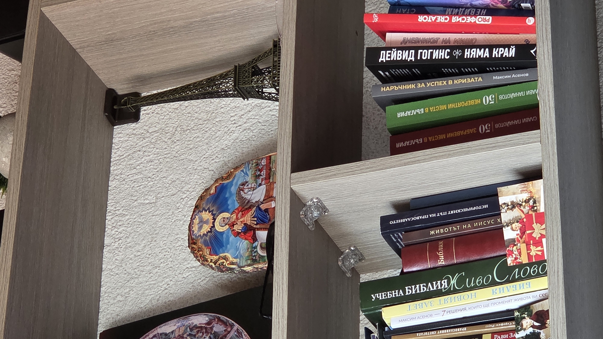 This image shows a bookshelf with various objects and books. On the left shelf, there's a decorative Eiffel Tower model and two decorative plates, one featuring religious iconography of a saint on horseback. The shelf is made of light-colored wood-like material, and the wall in the background is textured with a neutral color.
The right shelf is filled with books stacked vertically. The titles of some books are visible, mostly in Bulgarian, covering topics ranging from self-help ("Guide to Success in Crisis") to local attractions ("50 Incredible Places in Bulgaria"), and religious texts like the Bible and studies of Jesus Christ. There are two decorative, possibly crystal, bookends holding the books in place. The shelves are again of the same light-colored wood-like material.
