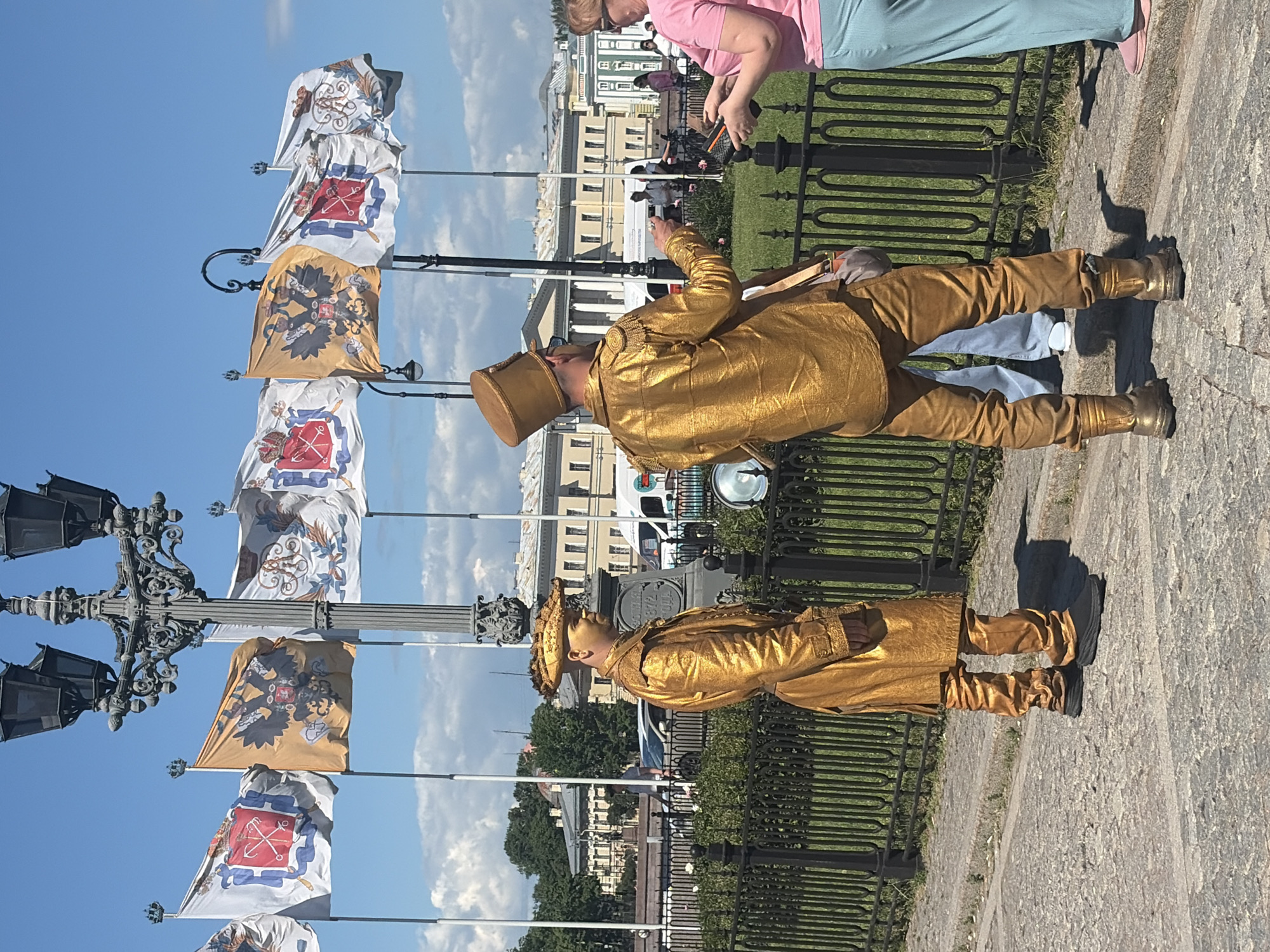 Two individuals dressed in full-body gold costumes, resembling historical military uniforms with ornate hats, stand on a cobblestone walkway in front of a black metal fence. One person, facing left, wears a long coat and pants, with a hat perched on their head. The other person, facing away from the camera, is also in a matching gold outfit, complete with a tall hat and a satchel slung over their shoulder. They appear to be interacting with a woman in a pink shirt and light blue pants, who is holding a device.
In the background, a series of flags with heraldic designs, including double-headed eagles and crowns, are displayed on poles against a bright blue sky with scattered clouds. Behind the flags, a grand building with a light-colored facade, classical architectural elements, and multiple windows is visible. Several vehicles, including cars and a white van with a logo, are present on a road in front of the building. Lush green trees and bushes form a line between the walkway and the road. The overall setting suggests a public square or promenade in a city known for its historical architecture, consistent with Saint Petersburg, Russia.