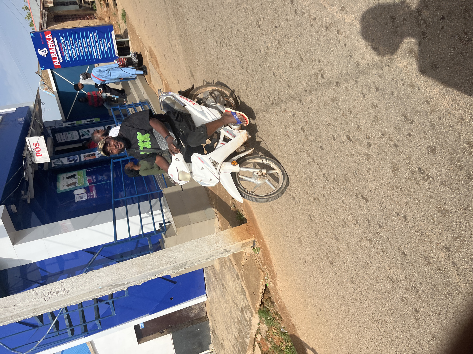 A man is riding a white motorcycle in front of a building with blue and white accents. He is wearing a black t-shirt with green lettering, black shorts, and flip-flops. The building has a sign that reads "POS" and another sign for "ALBARKA COMMUNICATIONS." There are other people in the background, some standing and some sitting. The scene appears to be in a commercial area with a paved road. The overall lighting suggests it is daytime. The location is Gura Top, Nigeria.