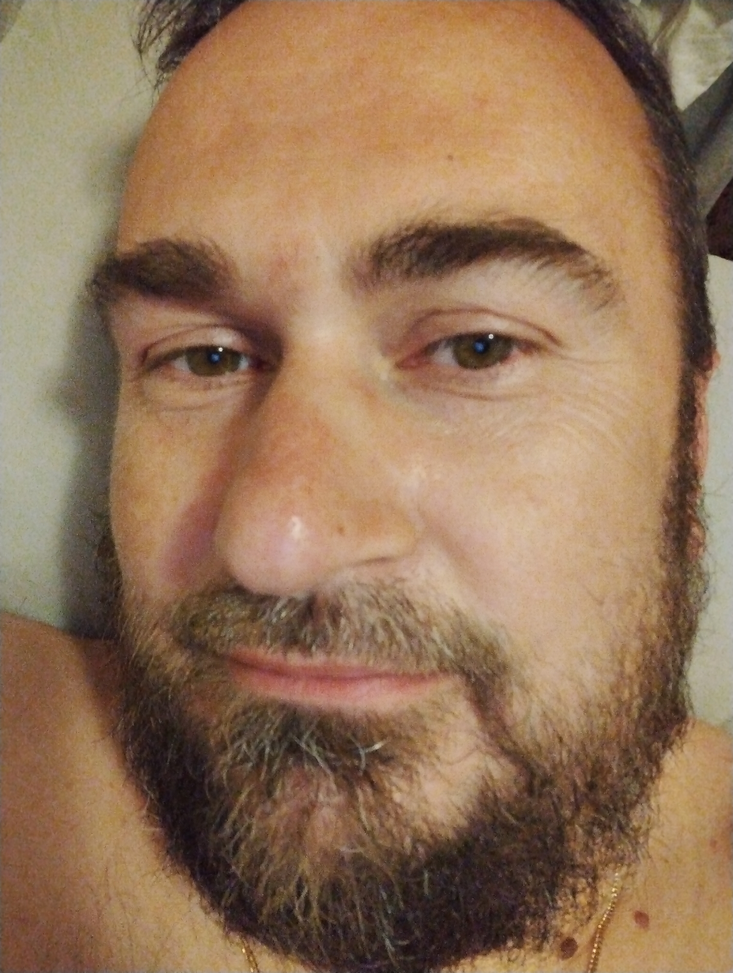This is a close-up, indoor, selfie-style portrait of a man's face, positioned vertically in the frame. The man has fair skin, brown eyes, and a beard. His eyebrows are dark and full, and his forehead shows some wrinkles. The background is out of focus and appears to be a textured wall or fabric. The lighting is soft and appears to be coming from the front. The image does not contain any discernible elements that place it in the city of Ширяево (ильинский), Russia.