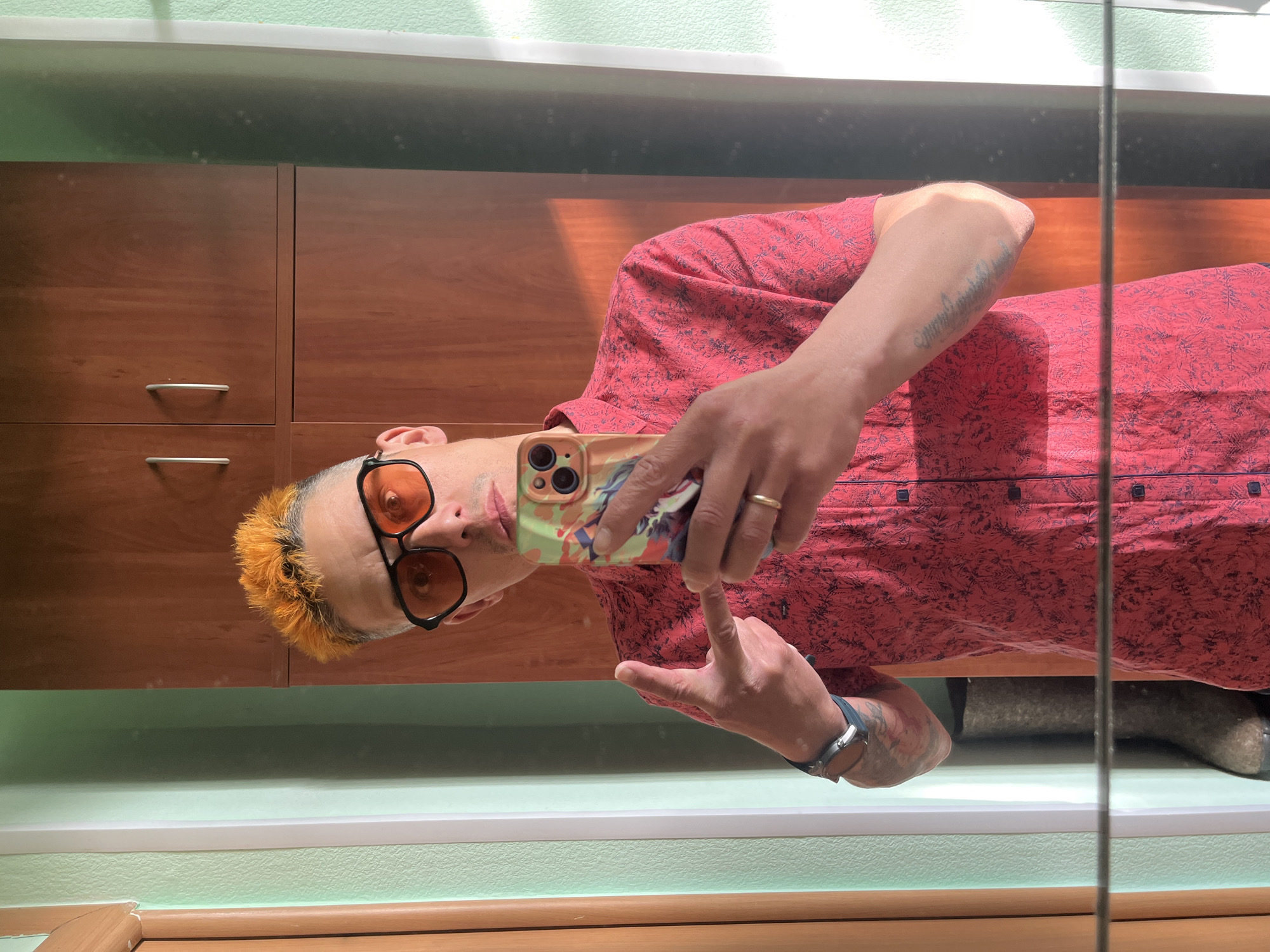 A person with dyed orange hair and wearing orange-tinted glasses is taking a selfie in a mirror. They are wearing a red patterned shirt and holding a phone with a patterned case. The background includes a wooden cabinet with chrome handles and a light green wall. The location is undetermined, and there is no confirmation that this is in Chaikovsky, Russia. The presence of shoes can be seen in the mirror in the lower right area of the image.