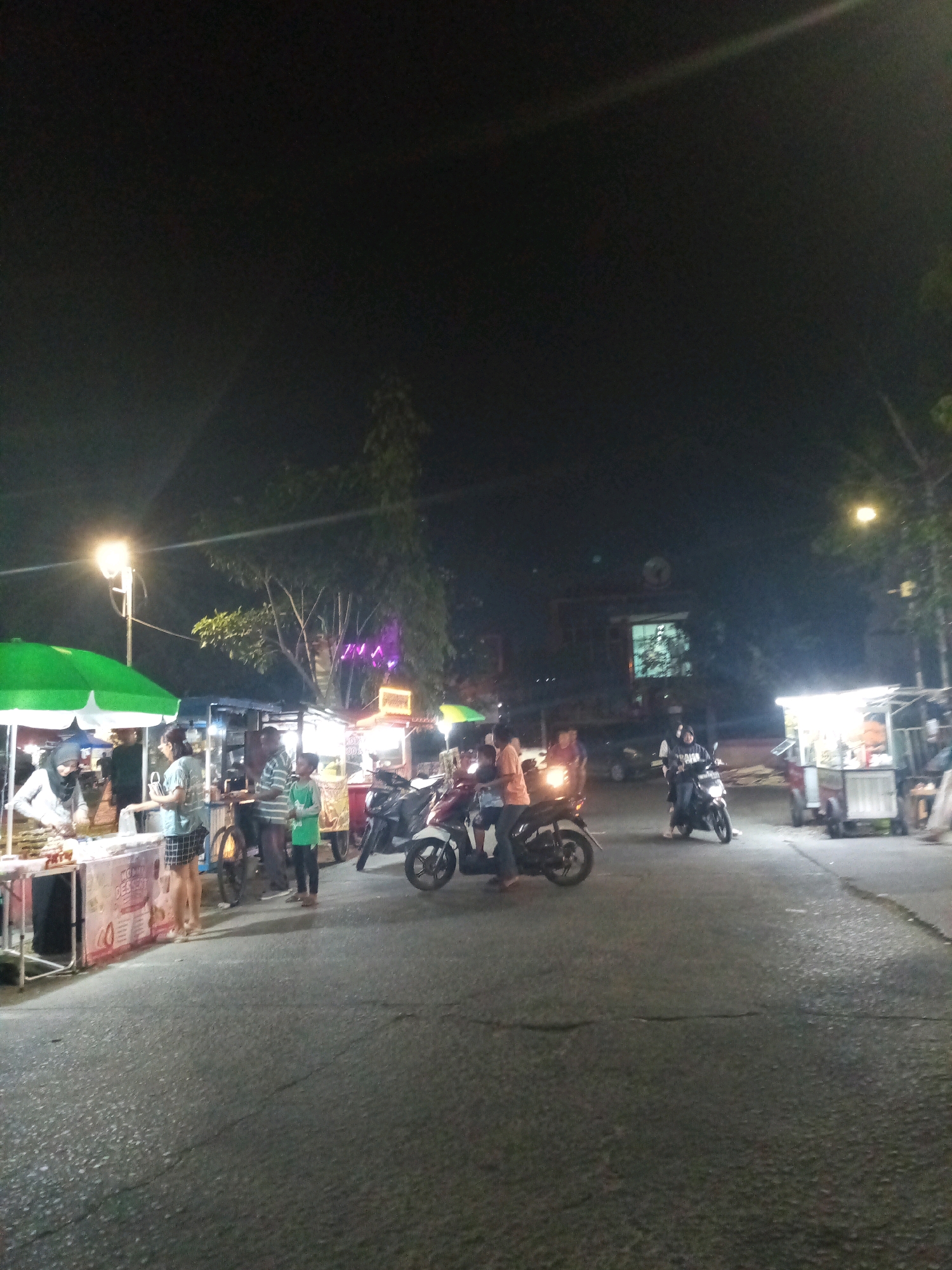 The image depicts an outdoor night scene, likely a street food market, in Bekasi, Indonesia. Several stalls with lights and umbrellas are visible, indicating commercial activity. People are present, some standing and interacting near the stalls, while others are on motorcycles, either parked or in motion. One stall has a green umbrella and appears to be selling food items. Another stall to the right has a bright white light illuminating it. The ground is asphalt and appears to be a road or pathway. The background is dark, suggesting it is nighttime, with some trees and buildings visible in the distance. The overall atmosphere suggests a lively evening market.