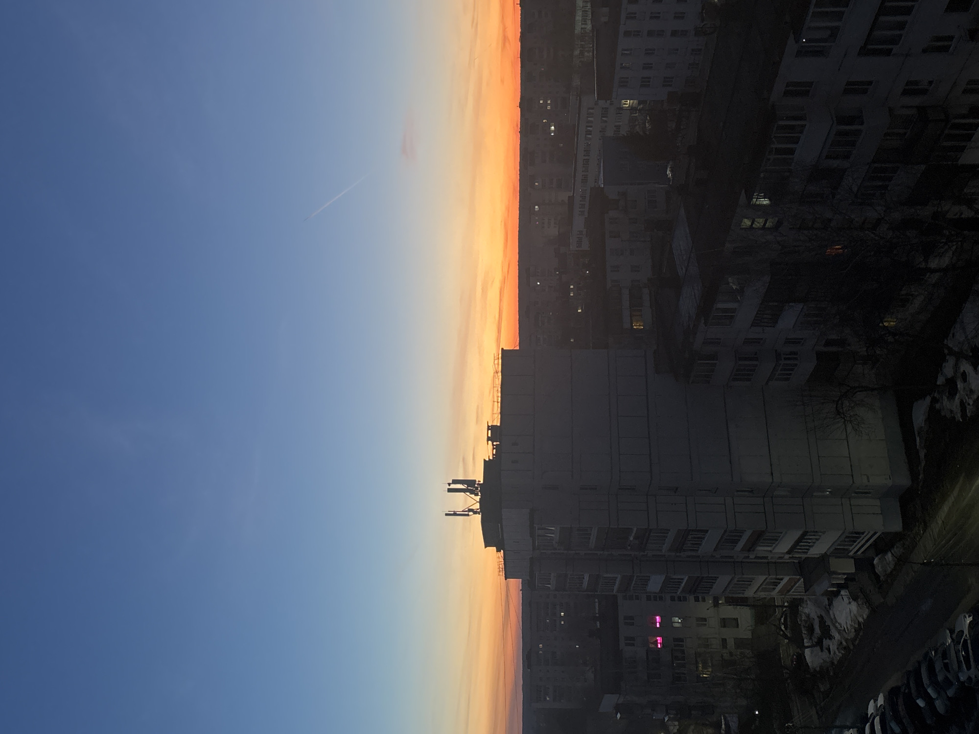The image depicts an urban landscape, likely in Chaikovsky, Russia, at dawn or dusk. The sky transitions from a deep blue to orange near the horizon, suggesting either sunrise or sunset. A contrail is visible in the blue sky. On the right side of the image, there are several multi-story residential buildings. Some windows are illuminated, indicating occupancy. One building closer to the foreground features antennas on its roof. The foreground shows dark shapes suggestive of cars parked below. The environment is lit by streetlights and the ambient light from the sky.