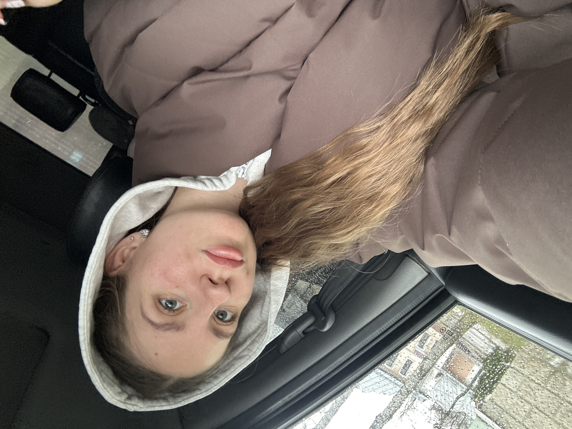 The image is taken from inside a car, with a young woman wearing a brown puffy jacket and a light gray hooded sweatshirt as the primary subject. Her long, light brown hair is visible on the right side of the frame. She is looking directly at the camera. Raindrops cover the car window, and the background shows a blurred view of a building and trees outside, which appears to be Klin, Russia, based on the building's visible Cyrillic sign reading "МАГАЗИНА" (SHOP). The interior of the car, including the seat and door, is dark gray.