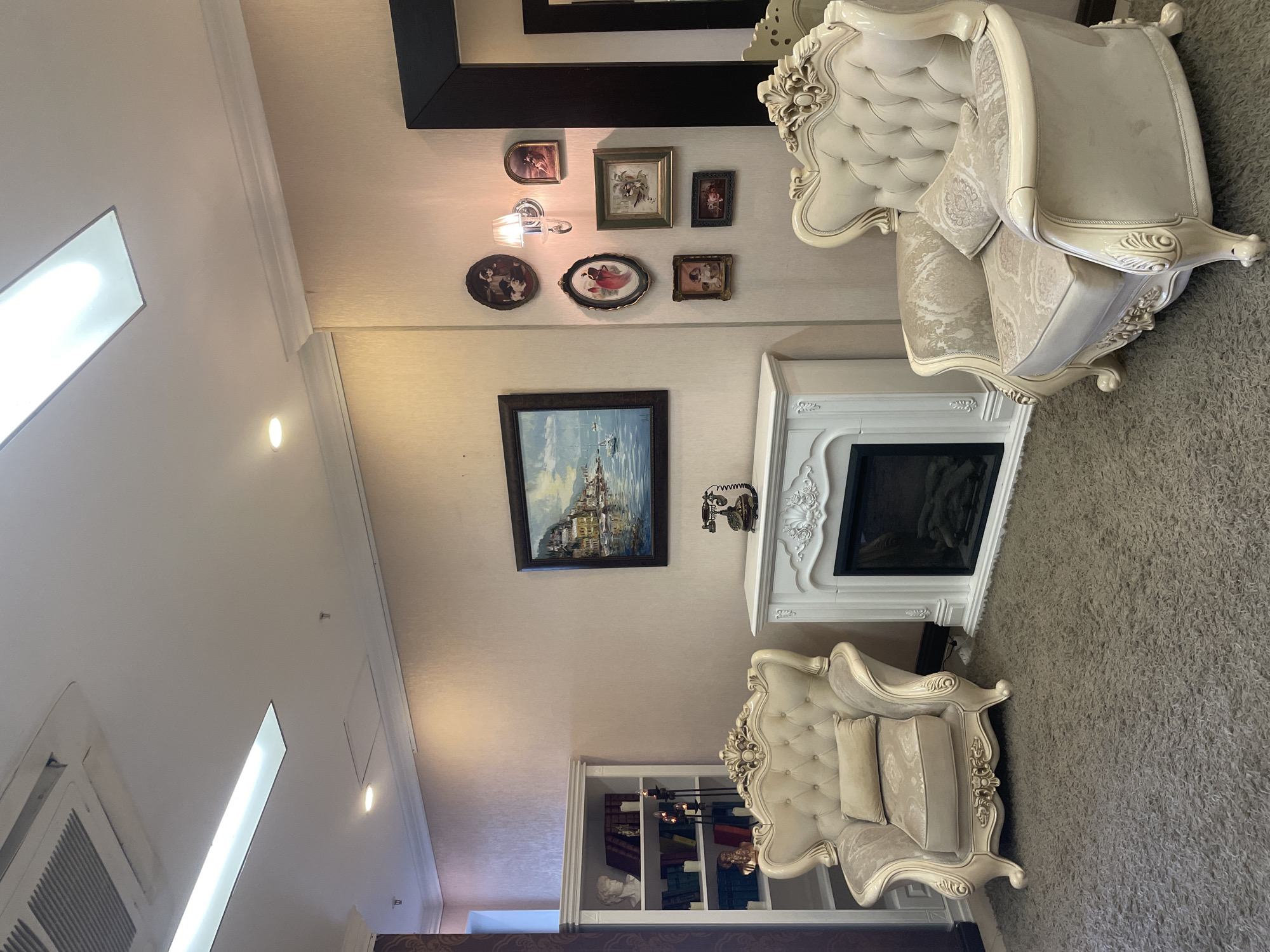 The image depicts an interior scene in Kazan, Russia, likely a decorative or showroom space. The room is furnished with two ornate, cream-colored upholstered armchairs positioned on a textured gray carpet. In the center of the frame, a white, decorative fireplace with intricate carvings stands between the chairs. Above the fireplace hangs a framed oil painting of a waterside town. To the left of the fireplace is a tall white bookshelf filled with books and decorative items, including busts and candles. To the right of the fireplace, a wall is adorned with a collection of framed portraits and a wall-mounted light fixture. A vintage rotary telephone rests on top of the fireplace. A large framed mirror is visible on the far right of the image. The ceiling features recessed lighting and an air conditioning unit. The overall ambiance suggests a classic or vintage aesthetic.