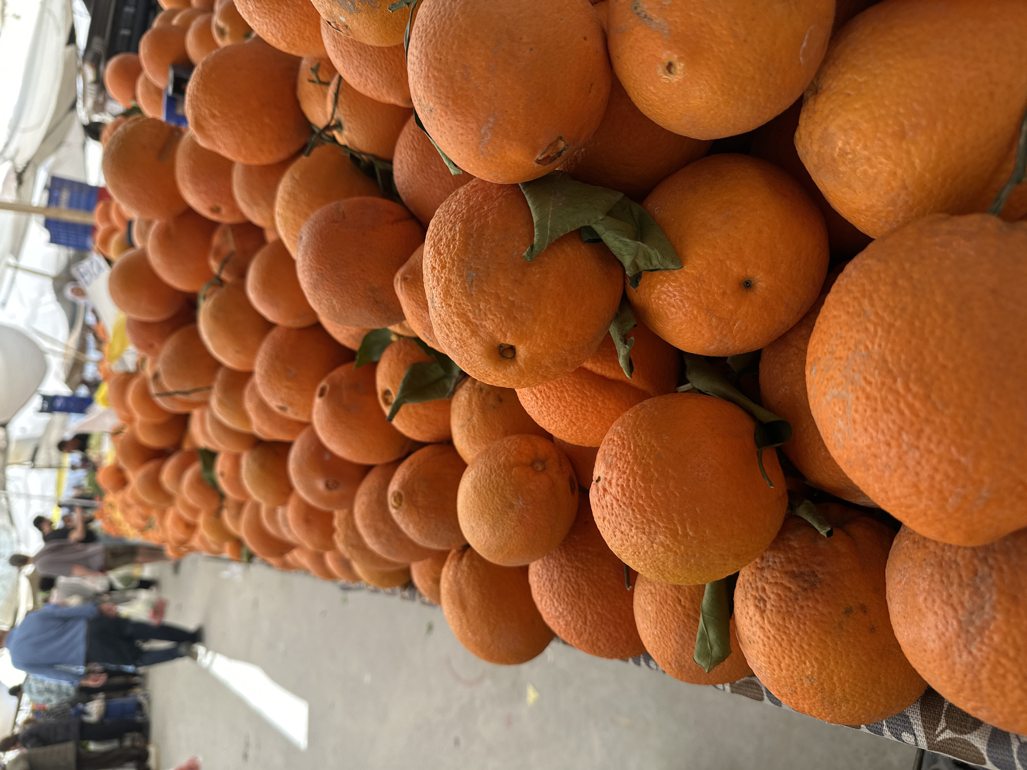 A close-up, slightly canted, outdoor shot shows a large number of oranges stacked at a market stall in Fethiye, Turkey. The oranges are densely packed together, creating a wall of orange color, and some have green leaves attached. The camera angle is from a low perspective, looking up at the stack of oranges. In the background, out of focus, are people walking in the market, along with the structures and coverings of market stalls. The ground is a gray concrete surface. The lighting appears natural and somewhat diffused.