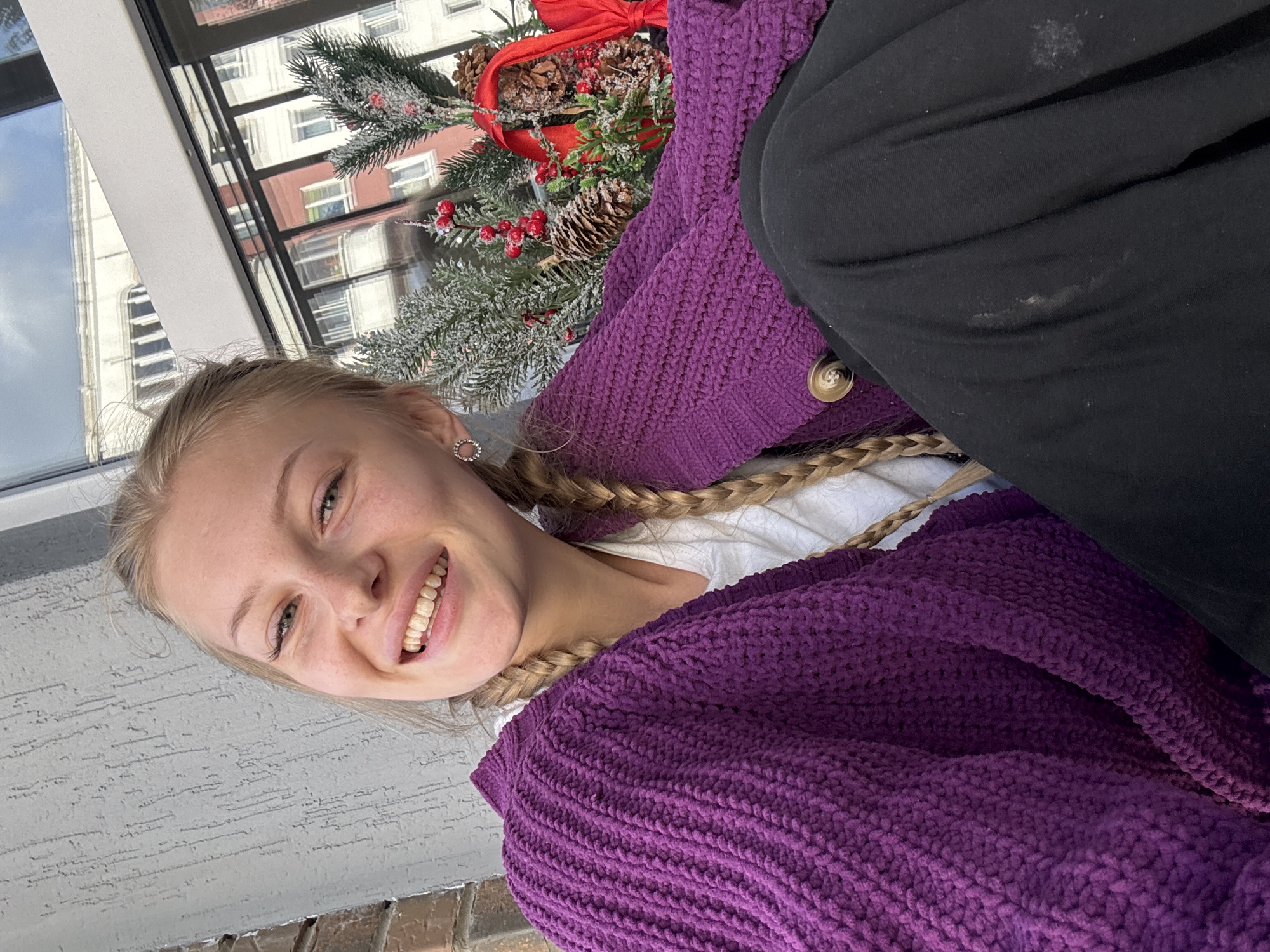 In Perm, Russia, a young blonde woman with braided hair and a bright smile is pictured outdoors. She is wearing a vibrant purple knitted cardigan over a white shirt and black pants. The woman is sitting near a brick wall and a window that reflects buildings and the sky. A small Christmas-themed decoration, featuring pine sprigs, pinecones, and red berries, is visible to her right. The overall scene suggests a casual, possibly festive, moment captured on camera.