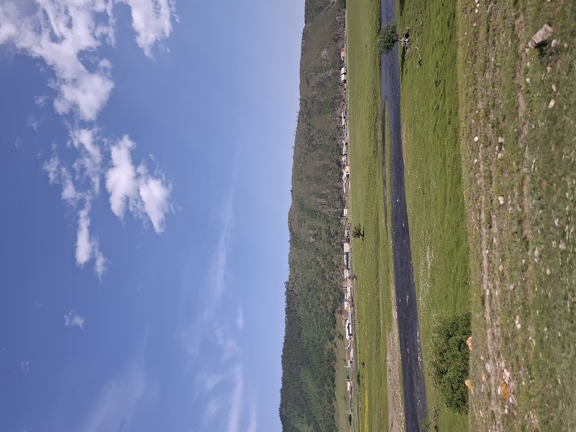 The image depicts a rural landscape under a bright blue sky with scattered white clouds. The scene is split vertically, with the left side dominated by the sky and a green, mountainous backdrop, while the right side features a river, a grassy field, and a dirt path.
In the mid-ground, a line of buildings, possibly houses or small structures, runs along the base of the mountain, suggesting a small village or settlement. The mountain itself is covered in green vegetation. A dark, narrow river flows through the middle of the frame, bordered by lush green fields. Small white flowers are visible in the grass near the river. The right side of the image shows a rough dirt path with scattered stones and patches of dry grass, indicating a less cultivated area.
The lighting suggests it is daytime, likely late morning or early afternoon, given the bright sunlight and clear skies. The weather appears to be fair and pleasant. The overall impression is one of a peaceful, natural setting in a remote location. No people or specific activities are directly visible, but the presence of the settlement implies human habitation. The location is identified as Bugul'deika, Russia.