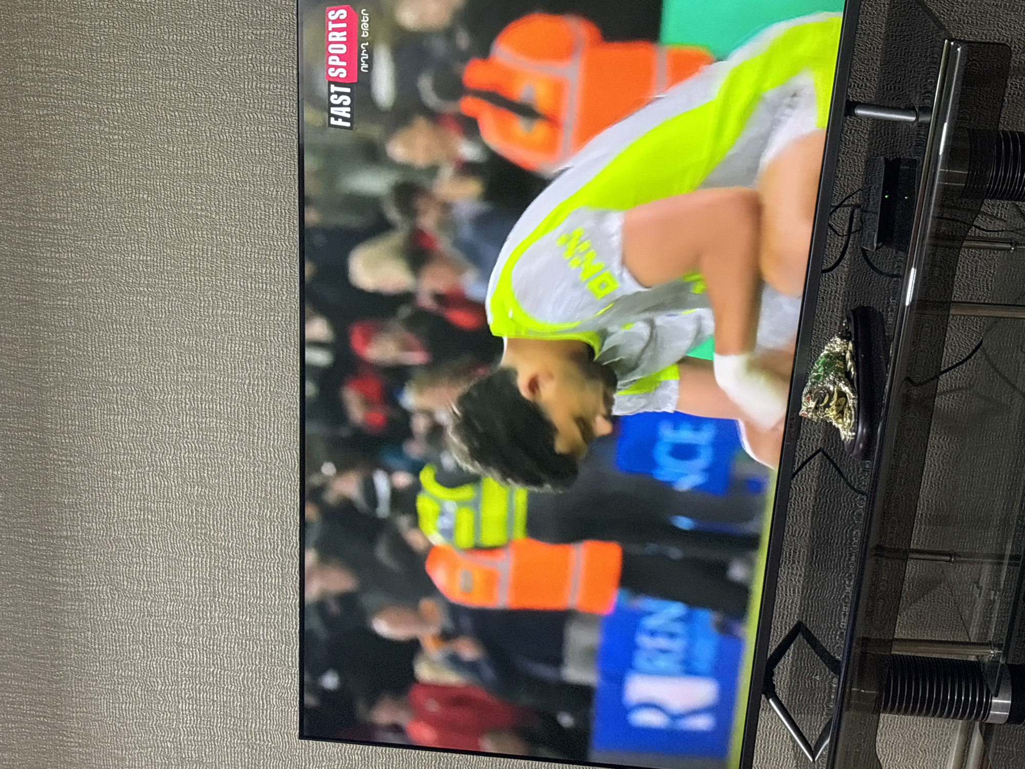 The image shows a television screen displaying a sporting event, specifically soccer. The "FAST SPORTS" logo and text in Armenian are visible in the upper left corner of the screen. The image is a close-up shot of a soccer player in a white and neon green jersey, leaning forward. The background shows a crowd of spectators and staff wearing orange vests and jackets, and there is a sign with the text "REN." The image is of a soccer match from city of Yerevan, Armenia.
