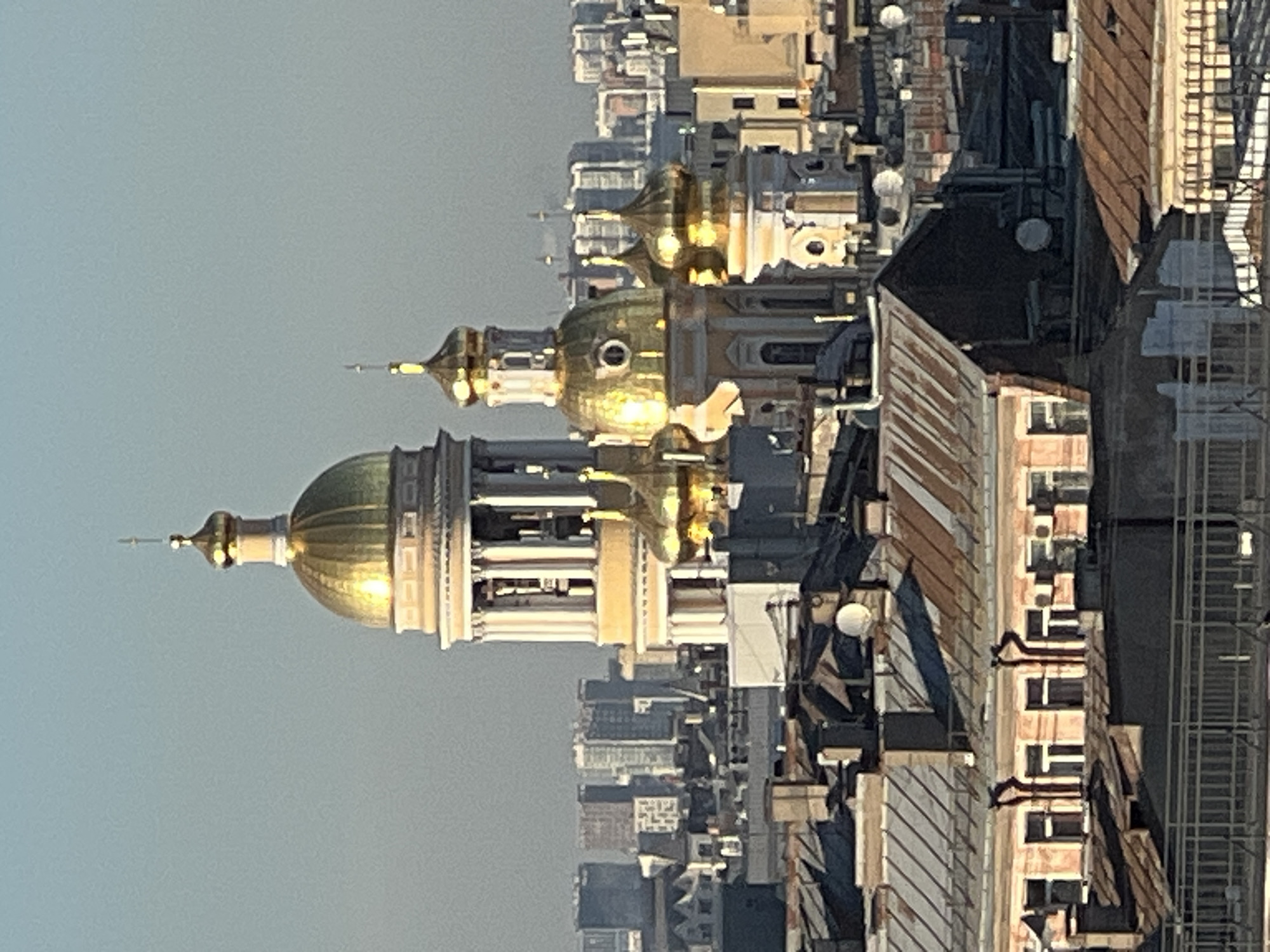 The image shows a cityscape in Saint Petersburg, Russia, focusing on the golden domes of Saint Isaac's Cathedral. The domes are the most prominent features, reflecting sunlight. In the background, a dense urban environment includes various buildings with differing heights and architectural styles. Rooftops are visible with details like satellite dishes, antennas, and varying materials. The sky is a pale, overcast color.