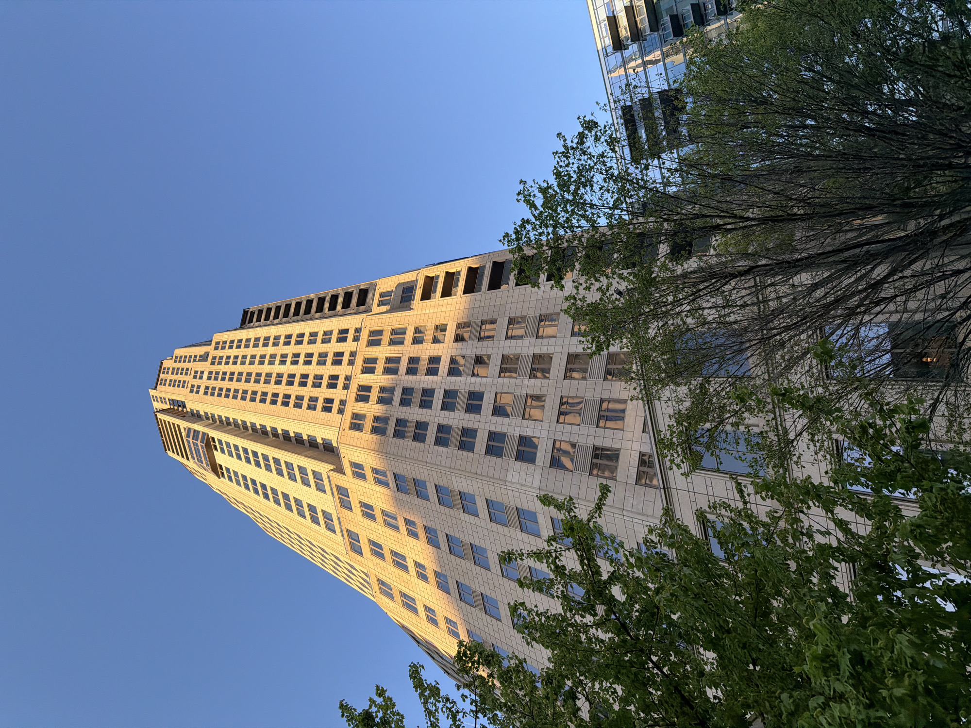 The image captures a low-angle view of a skyscraper in Atlanta, USA, juxtaposed against a clear blue sky and the foliage of trees. The building has a tan or beige facade with a grid of rectangular windows, reflecting sunlight. The architecture is modern, with a layered design at the top. To the right, a neighboring building with a glass facade and reflective windows can be partially seen, obscured by green tree branches with leaves. The foreground features tree branches, some bare and others with green leaves, adding a natural element to the urban scene.