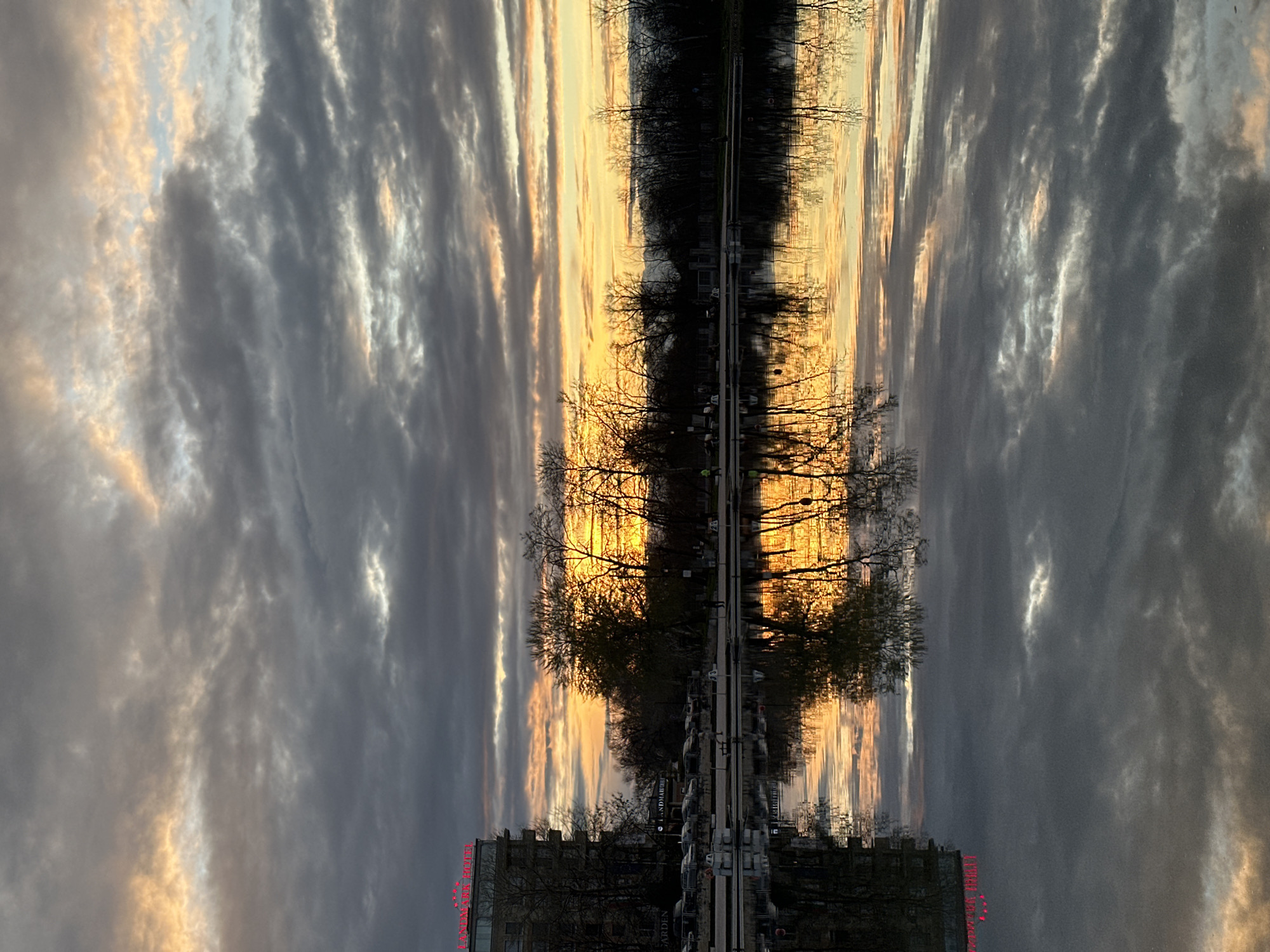 The image depicts a twilight scene in Plovdiv, Bulgaria. The building at the bottom appears to have neon lights that say "LANDMARK RIVER GARDEN." This building and the row of trees in front of it are reflected in the water. Above the tree line is a sunset reflecting off the clouds.