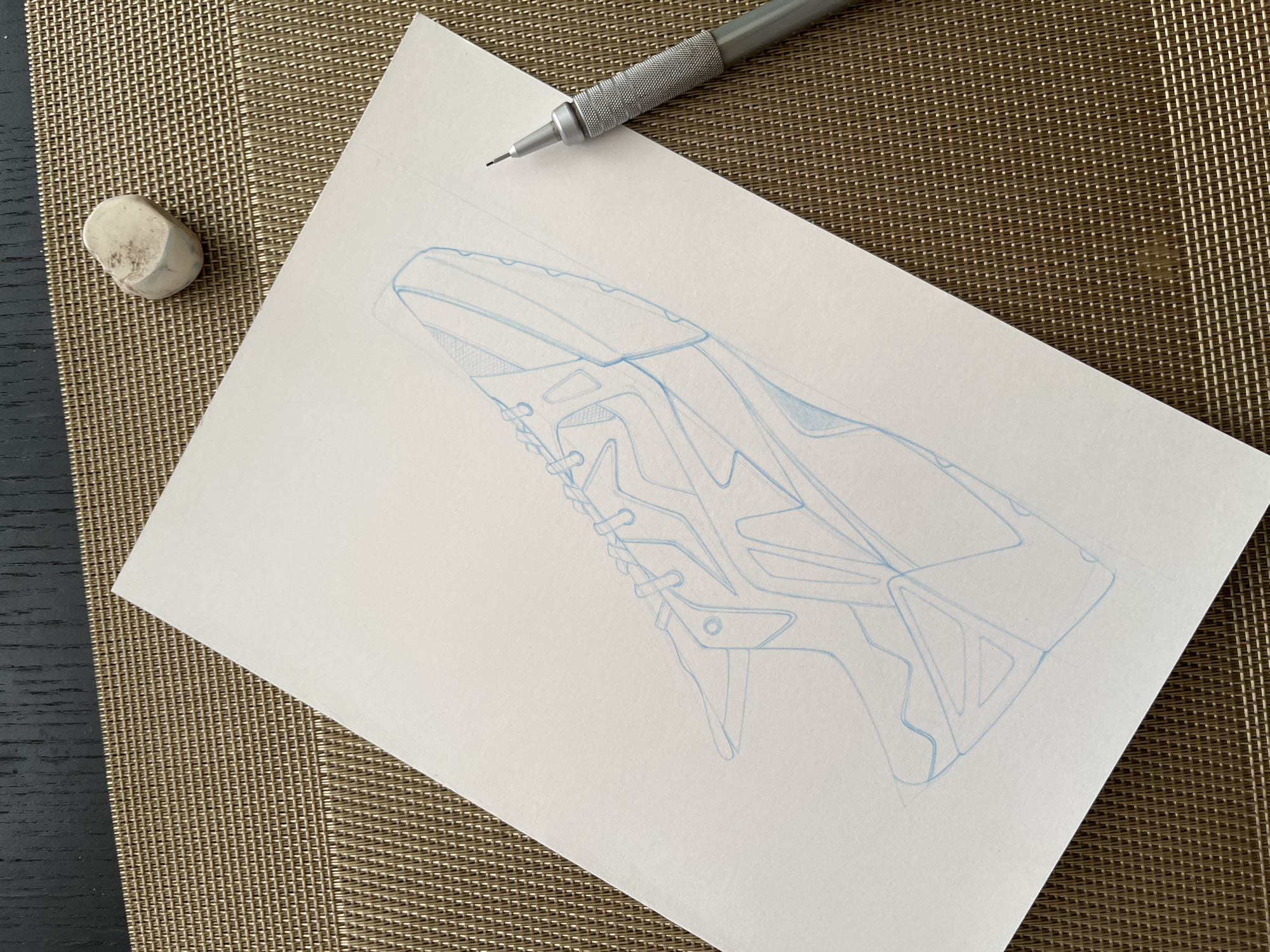 The media file presents an overhead view of a drawing setup, likely on a desk or table, in an indoor setting in Krasnoyarsk, Russia. The central focus is a white sheet of paper featuring a detailed technical sketch of what appears to be a modern or futuristic shoe or boot, drawn with precise light blue lines. The design incorporates angular shapes, textured sections, and structural elements typical of product or industrial design.
A silver mechanical pencil with a distinct knurled grip rests diagonally across the top right corner of the paper, its lead extended, suggesting it has been recently used. In the top left corner, a small, off-white, and visibly worn eraser is positioned.
The paper and drawing implements are situated on a golden-brown woven placemat, which covers the majority of the surface. A dark, possibly wooden or laminate, table surface is visible along the left and bottom edges of the frame, beneath the placemat. The lighting appears bright and even, indicating daytime. No individuals are present in the image, and there is no visible text on any of the objects. The scene conveys an ongoing creative or design activity.