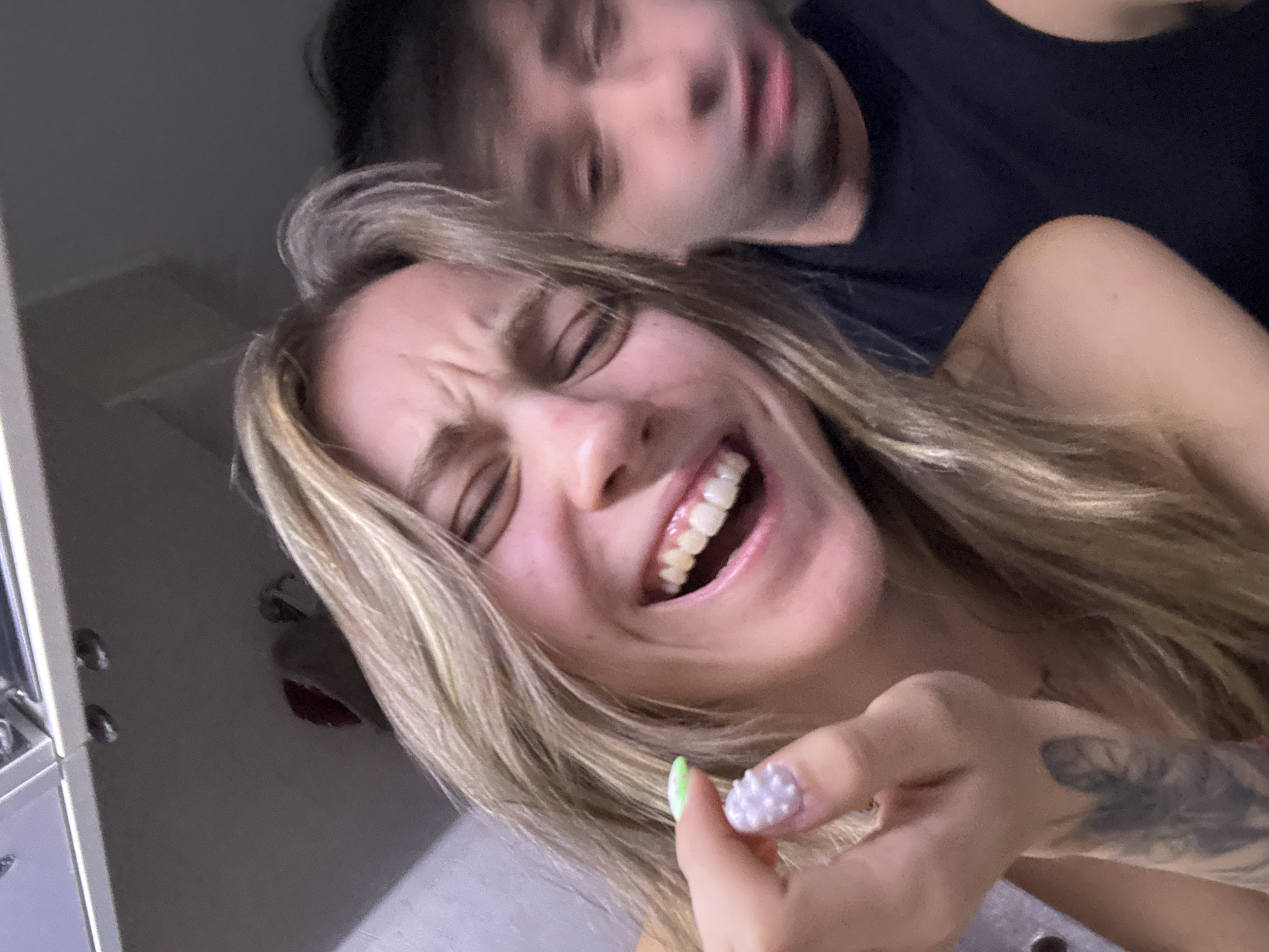 This media file is a close-up, selfie-style photo of two people. The foreground subject is a woman with blonde hair and fair skin, captured mid-laugh with her eyes squeezed shut and her mouth open, revealing her teeth. She is wearing a black top, and her right hand is visible, holding a small white object. Her fingernails are adorned with decorative pearls on one finger and a light green polish on another. A tattoo is visible on her forearm. Behind her, slightly out of focus, is a man with dark hair and fair skin, also looking towards the camera with his mouth slightly open. The overall mood appears joyful and playful.
The scene is indoors, with a blurry background that suggests a domestic setting, possibly a bedroom or living area, with some reflective surfaces and indistinct objects. There are no clear indications of the time of day or weather conditions. The location context provided is Moscow, Russia. There is no visible text in the image.