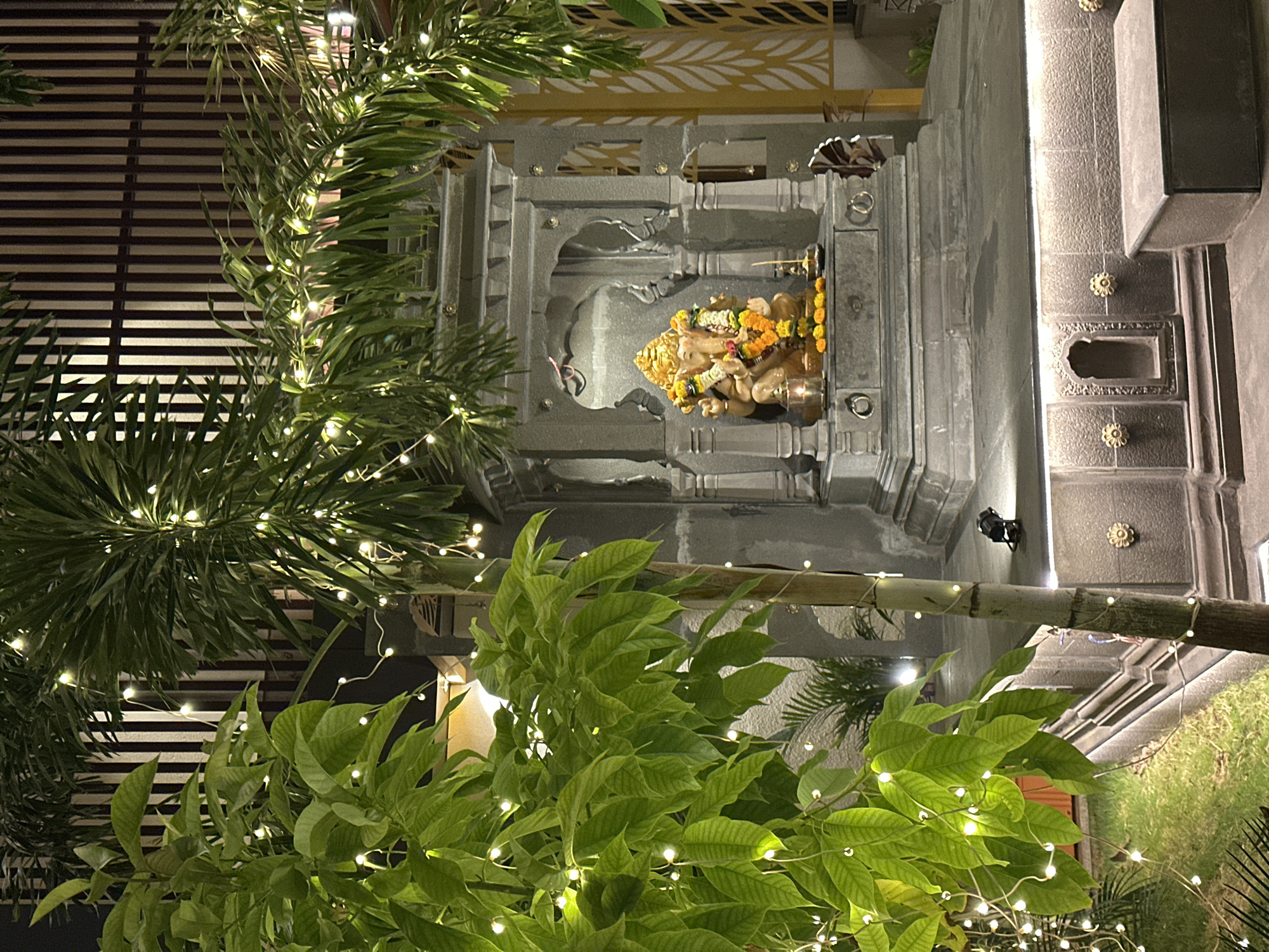 A statue of Ganesha, adorned with yellow flowers, is visible in an ornate gray niche in Kalamb, India. The statue appears to be made of a golden-colored material, possibly bronze. The niche is elaborately designed with multiple layers and pillars, all finished in gray. A dark grille or screen is visible in the background. The foreground is filled with green plants with bright green leaves, intertwined with strings of small white lights. These lights add a festive atmosphere to the scene. The image is shot from a low angle, looking upwards, emphasizing the height of the niche and the surrounding foliage.
