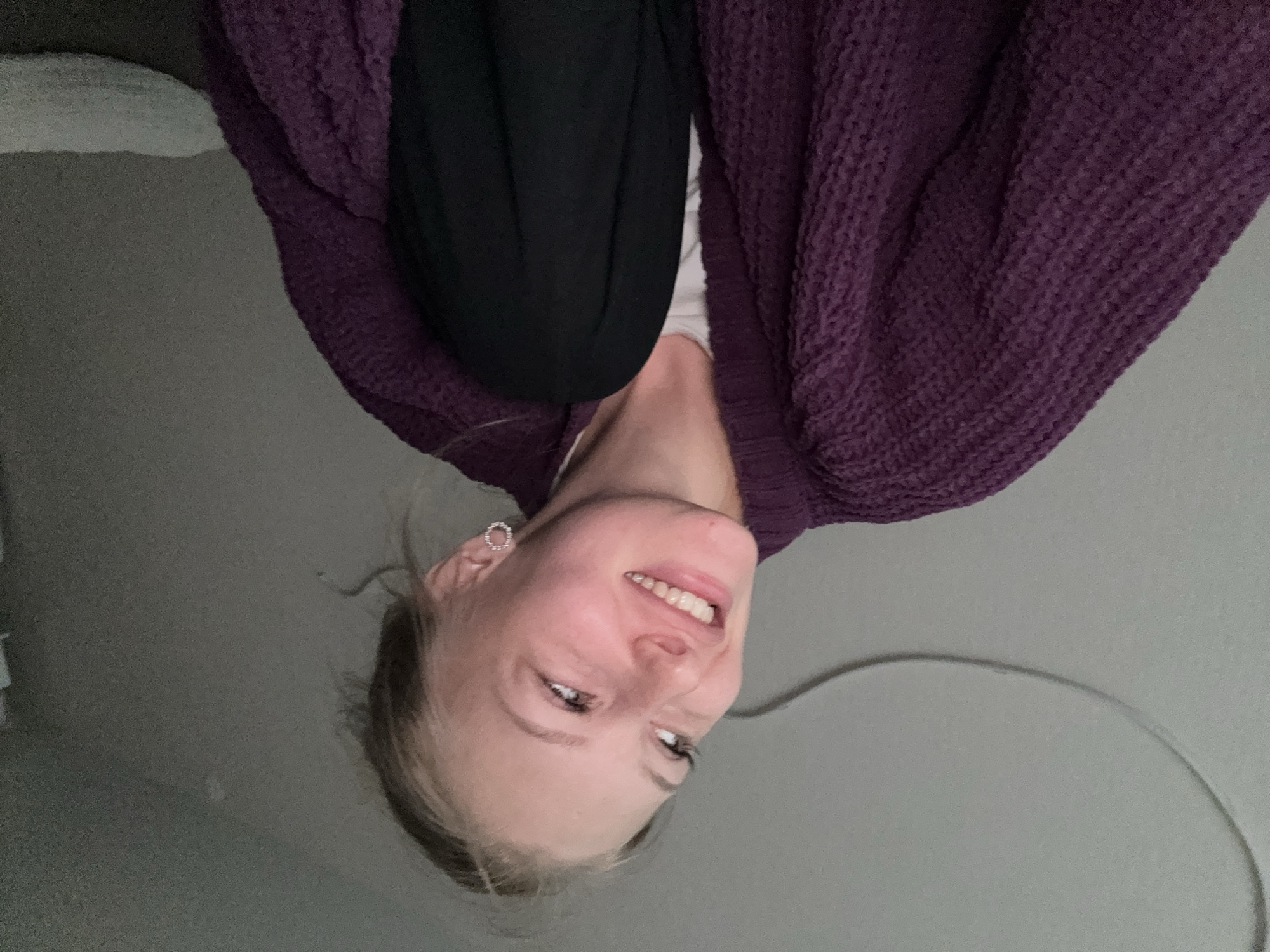 A young woman with blonde hair is shown from the chest up. She is wearing a dark purple, textured cardigan over a white shirt and black pants. She has a circular earring in her right ear. She is looking up and to her left with a smile. The background is a plain, grey wall. No information about the location in Perm, Russia is present in the image.