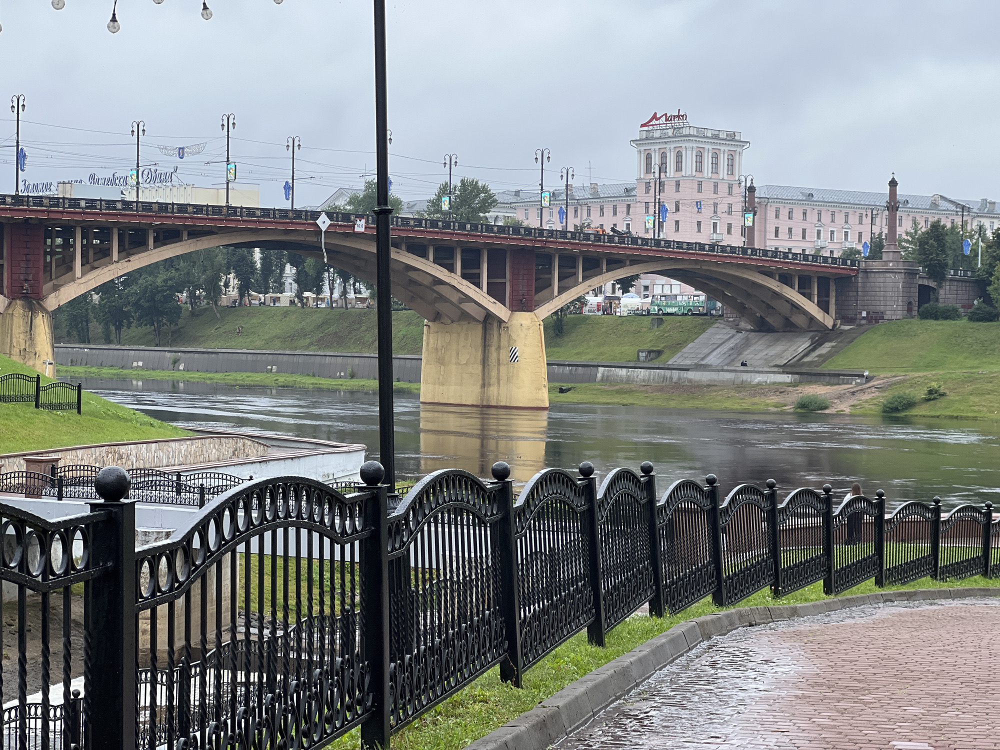 The image captures a view of the Dvina River in Vitebsk, Belarus, on a cloudy day. In the foreground, a black ornate metal fence runs along a wet brick walkway and a strip of green grass. Beyond the fence, the Dvina River flows, its surface reflecting the overcast sky. A large arched bridge, with red and yellow accents, spans the river. On the far bank, a green grassy slope leads up to a long, pale pink building topped with a red "Marko" sign. The building has many windows and architectural details. Several streetlights and wires are visible above the bridge. On the grassy bank beneath the bridge, some white tents and a green bus are present, suggesting an event or market. The overall atmosphere is muted due to the weather.