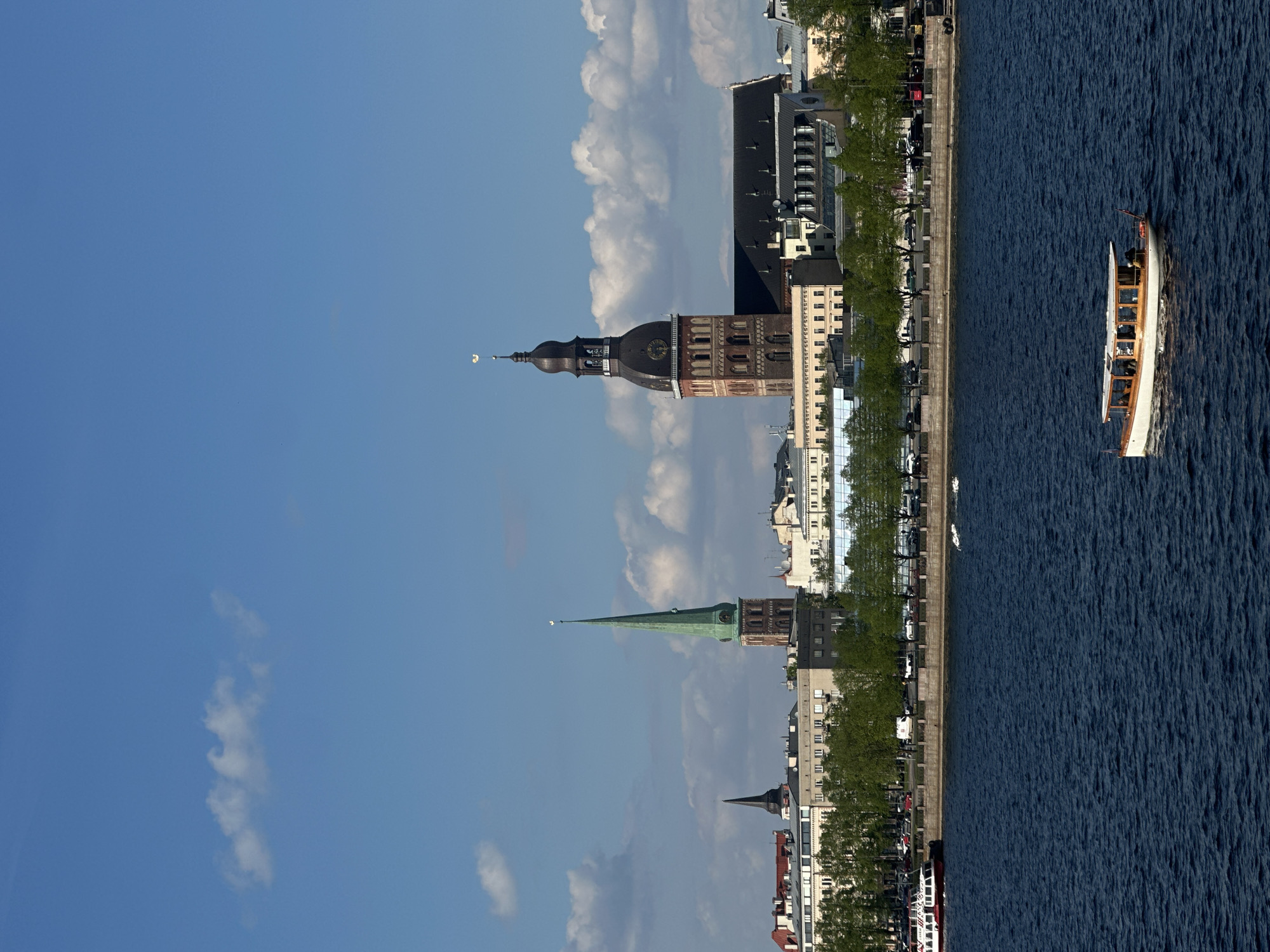 The image shows the cityscape of Riga, Latvia along the Daugava River. The spires of Riga Cathedral, St. Peter's Church, and St. James's Cathedral are visible against a blue sky with scattered clouds. A passenger boat travels along the river, parallel to the embankment with trees and buildings. Cars are parked along the riverside. The scene is bathed in sunlight.