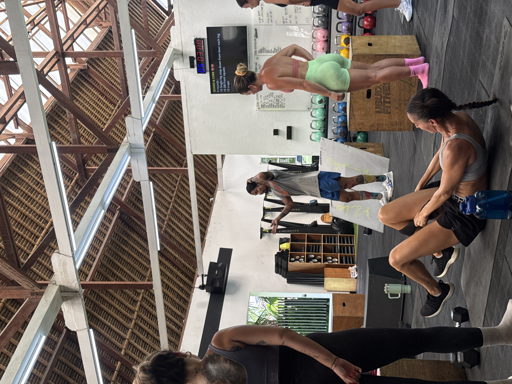 Here is a matter-of-fact description of the image:
The image captures a fitness class in progress, possibly at a gym or fitness center located in the Ubud District of Indonesia, as suggested by the text on a nearby box. The space has a high, exposed roof structure made of wood and supports, typical of architecture found in Ubud.
Several individuals are present. One woman is performing an exercise, possibly step downs, on a wooden box. Another woman, who is braided, is sitting on the floor, resting or recovering, with a water bottle beside her. Others are also exercising in the background.
Training equipment such as kettlebells and dumbbells are visible on shelves and the floor, indicating a crossfit gym. There's a digital clock displaying the time "05:03:00" and a whiteboard with training instructions listed, including "QUADS & CORE" and specific exercises. An air conditioner is also visible.