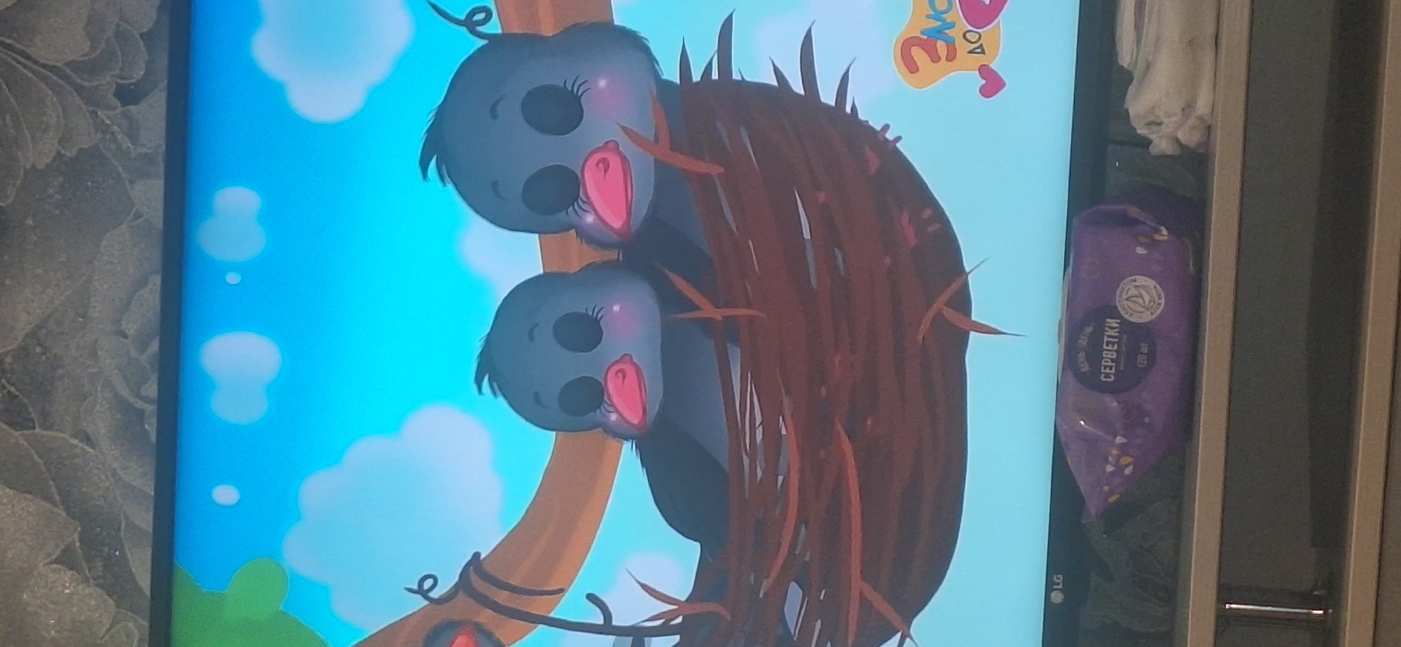 The image displays a television screen showing an animated scene of two cartoon birds in a nest on a tree branch, set against a bright blue sky with clouds. Below the television, on a shelf, is a purple package of wet wipes labeled in Cyrillic script, and to its right are folded white cloths. The television itself has an "LG" logo visible on its bottom left corner. The scene is captured from a slightly elevated angle, looking down at the television and the shelf. The background behind the television is a textured wallpaper. The image does not contain any specific location markers within the city of Bila Tserkva, Ukraine.