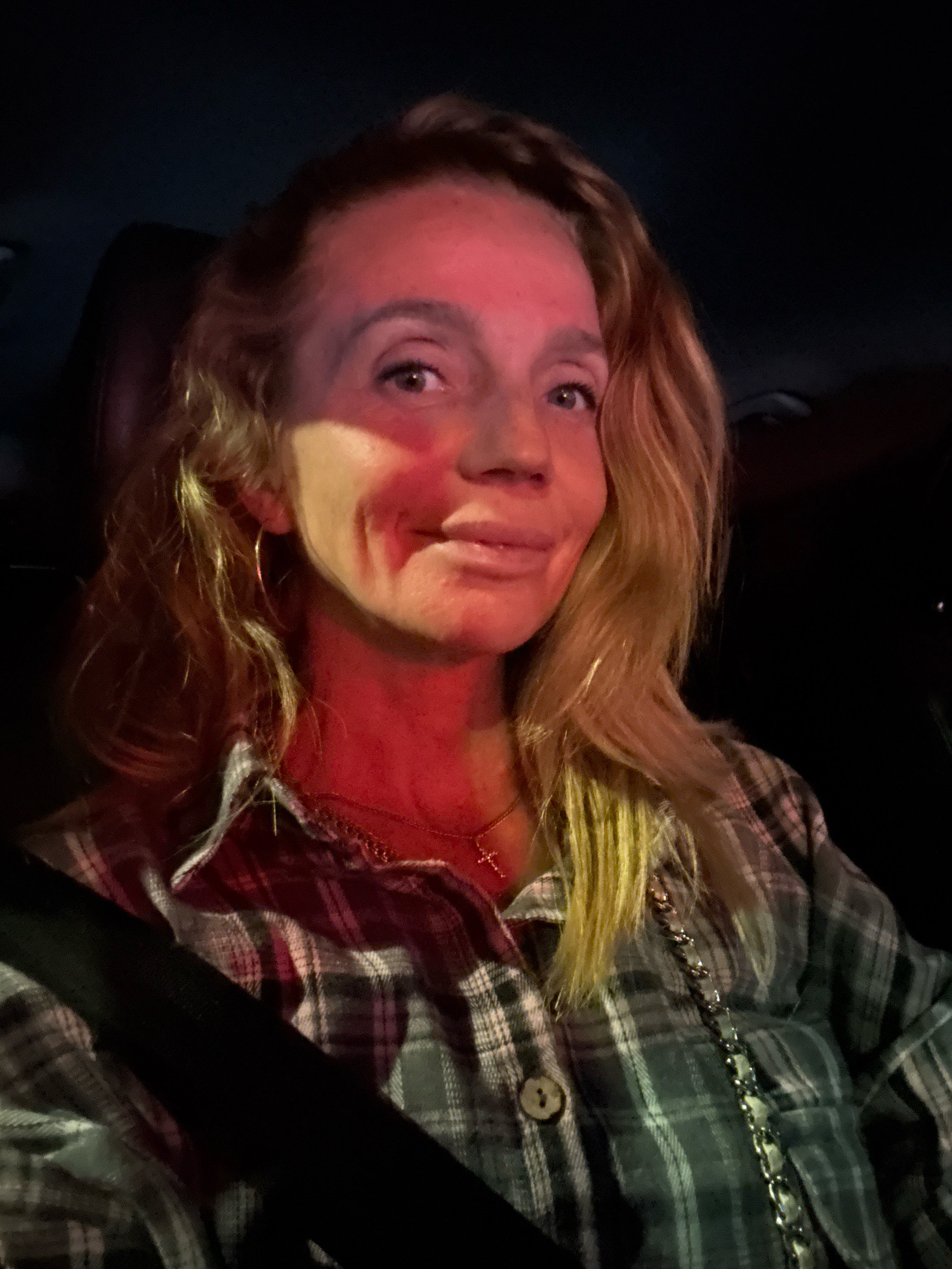 In a dimly lit environment, likely a car interior given the visible seatbelt and dark background, a woman with blonde, wavy hair is captured in a close-up, frontal shot. Her face, illuminated by a strong red light source originating from her left, displays a subtle smile and her eyes are looking directly at the camera. She is wearing a plaid shirt with a visible button and a necklace with a cross pendant. The lighting creates dramatic shadows and highlights, particularly across her face and hair. The scene does not contain any identifiable landmarks or context to suggest a specific location within Saint Petersburg, Russia, or any other city.