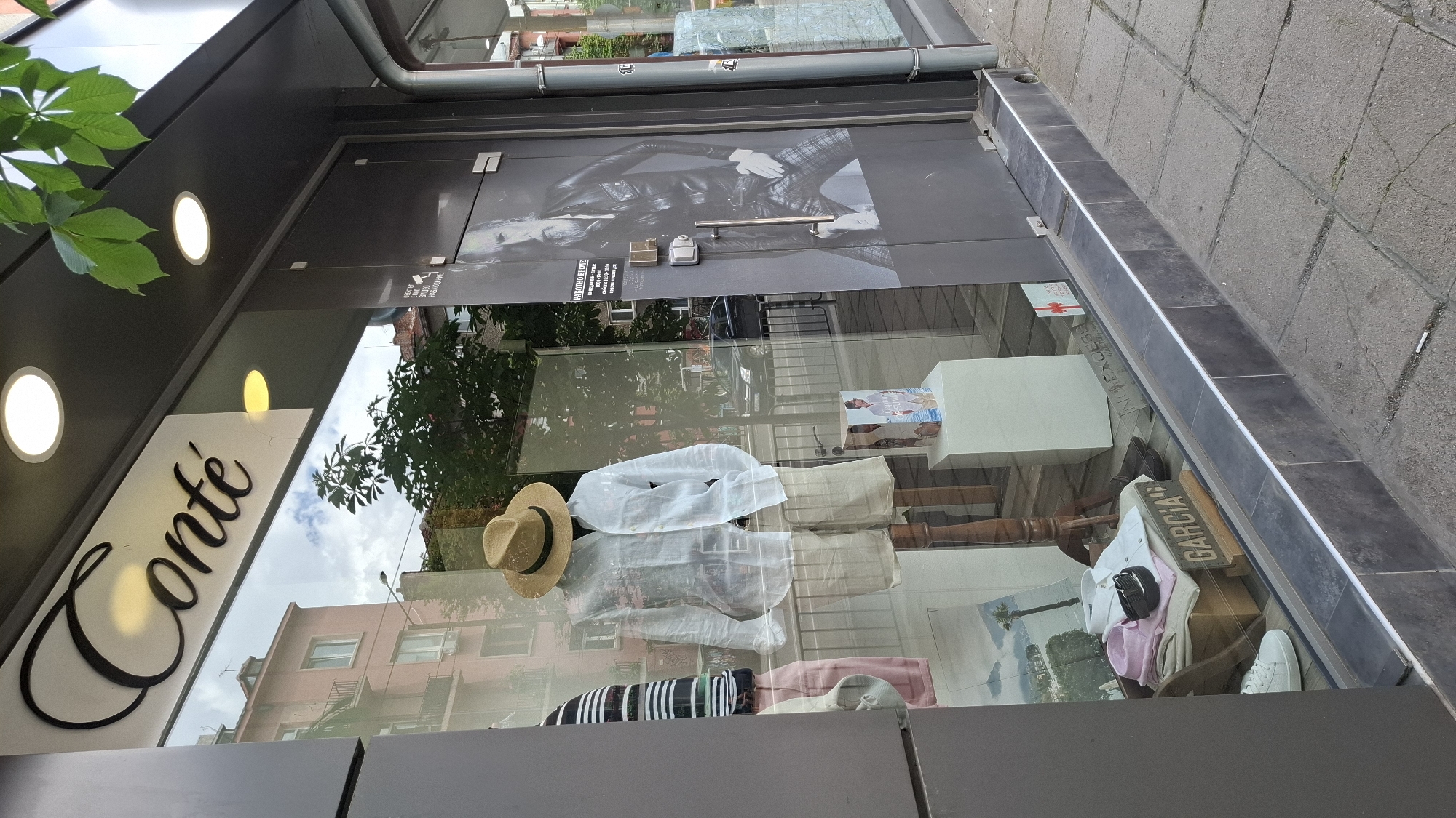 This media file captures a street-level view of a clothing store's storefront in Sofia, Bulgaria. The primary subjects are the store's display window, filled with clothing items and mannequins, and the exterior of the building.
The store features a large glass display window showcasing various apparel, including shirts, a straw hat, and a striped scarf or garment. Several mannequins are arranged to display the clothing. One mannequin prominently features a white shirt and a straw hat. Another display shows a pink sweater and a white shirt. A wooden sign with the word "GARCIA" is visible in the display.
Above the display window, the store's facade is visible, with a prominent sign featuring the word "Conte" in a stylized script, illuminated by a yellow light. Two circular white lights are also integrated into the facade above the "Conte" sign. The store appears to be a boutique or fashion retailer.
The reflection in the glass display window provides context about the surroundings. It shows buildings across the street, trees, and a cloudy sky, suggesting it's daytime. The weather appears to be overcast. The sidewalk in front of the store is paved with stone tiles.
Notable details include the signage of the store ("Conte" and "GARCIA"), the arrangement of clothing items for display, and the reflections indicating the urban environment of Sofia. The overall impression is of a commercial establishment in an urban setting, with a focus on fashion merchandising. No specific activity or event is taking place beyond the store being open for business.