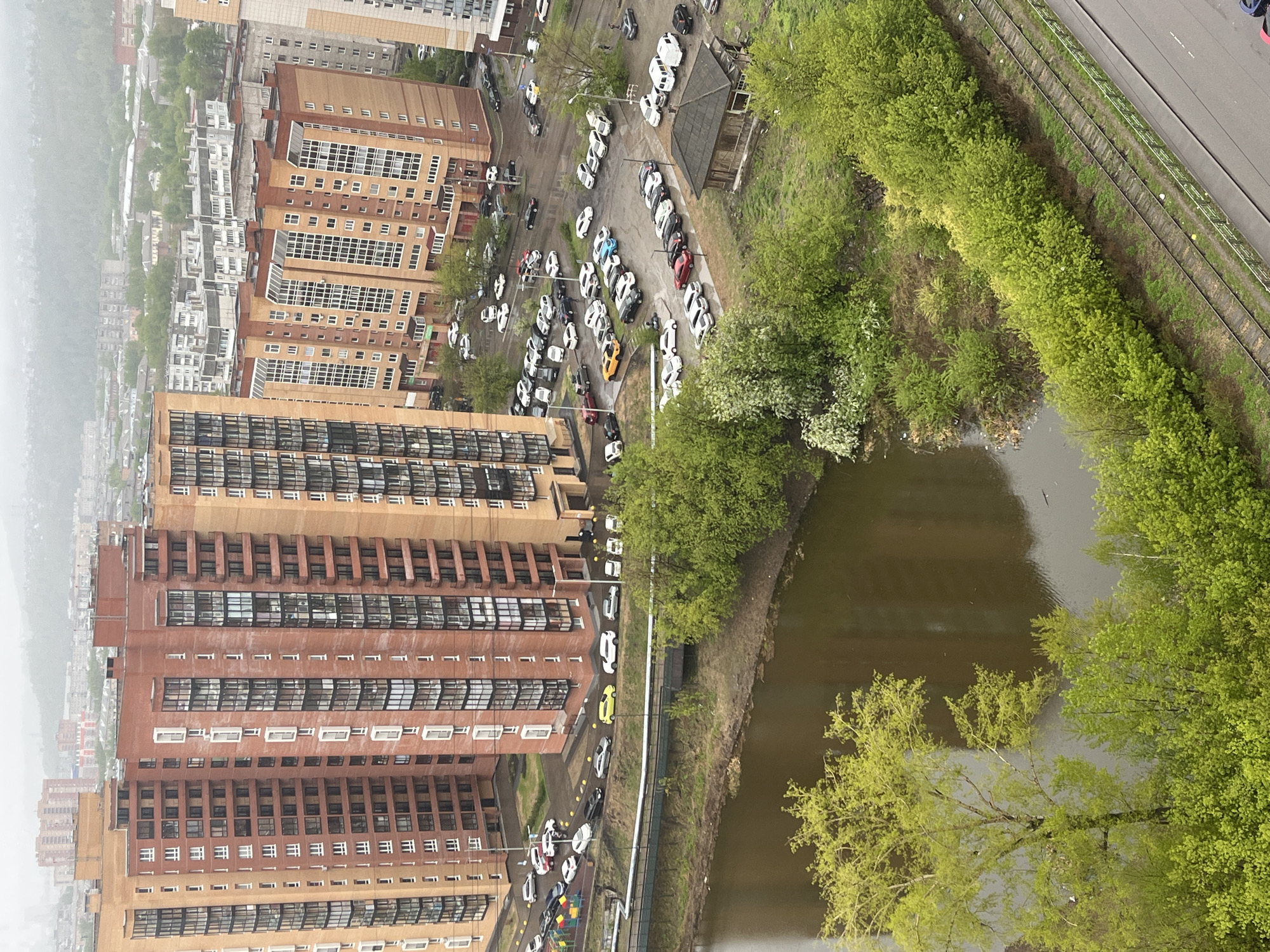 The image captures an elevated view of an urban landscape, likely taken from a high-rise building. It's daytime, and the lighting suggests overcast conditions. The scene includes multi-story buildings with brick and beige exteriors, situated near a body of water, possibly a canal or river.
To the right, a line of dense green trees follows the water's edge. A road and what appear to be tram tracks run parallel to the trees.
To the left, buildings with residential or office spaces are closely packed. In the central area between the buildings and the water, there is a parking area filled with numerous parked cars, mostly white, alongside a few other colored vehicles. There are some small structures or outbuildings near the parking lot.
Overall, the image conveys a typical urban setting with a mix of buildings, roads, transportation infrastructure, and natural elements. There are no visible people in the image.