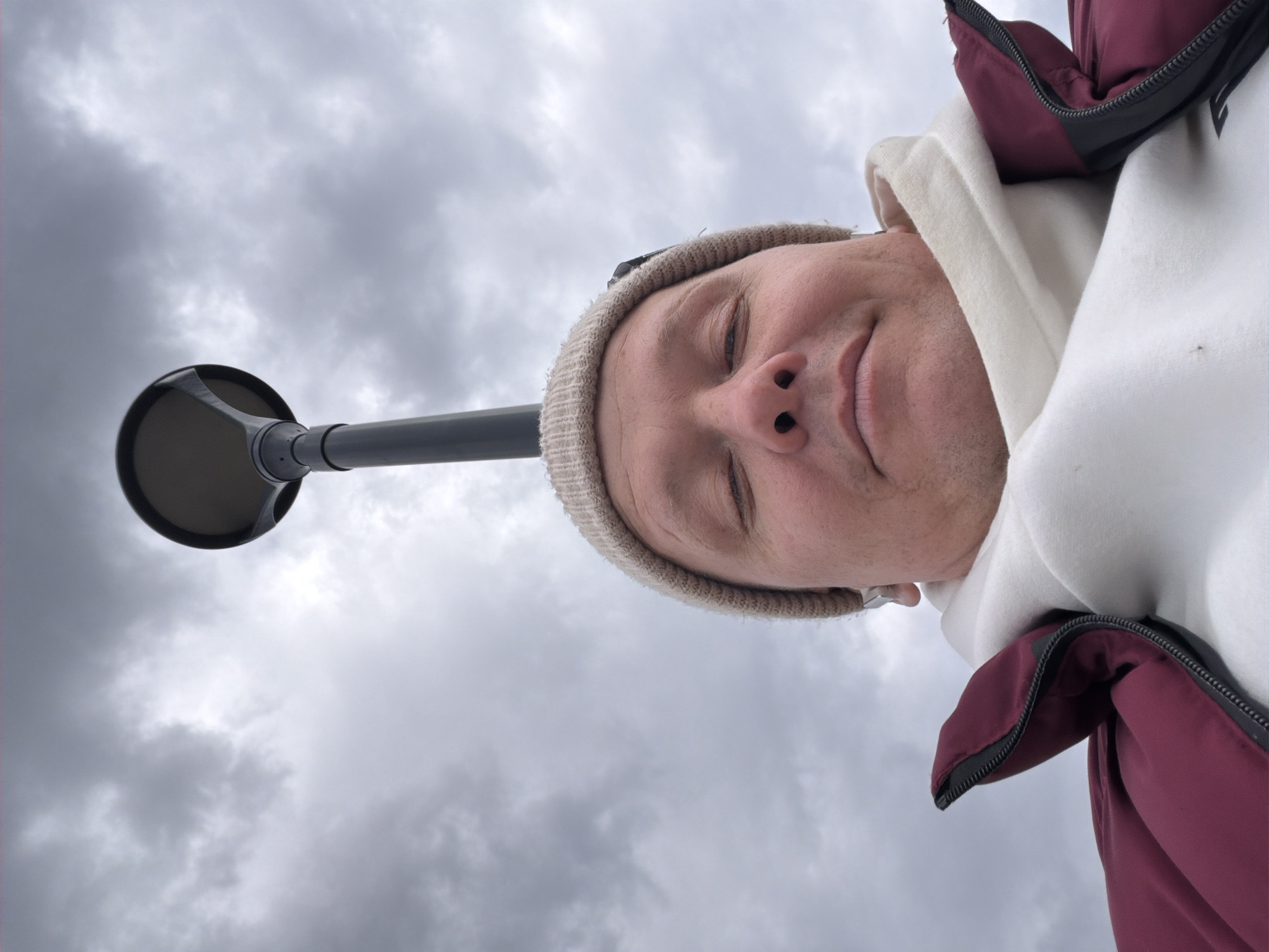 A low-angle shot, oriented as if portrait, captures a man looking upwards at the camera against an overcast sky. He is wearing a light-colored knit beanie, a white hoodie, and a dark red (burgundy) jacket with a black zipper. His eyes are partially closed, and a subtle, relaxed smile suggests a feeling of contentment or peacefulness. To the left, a grey pole with a dark, circular street lamp fixture extends into the frame. The background consists entirely of a cloudy, grey sky, indicating an outdoor, daytime setting with no direct sunlight. The scene likely takes place in a public area in Chaikovsky, Russia. There is no visible text.